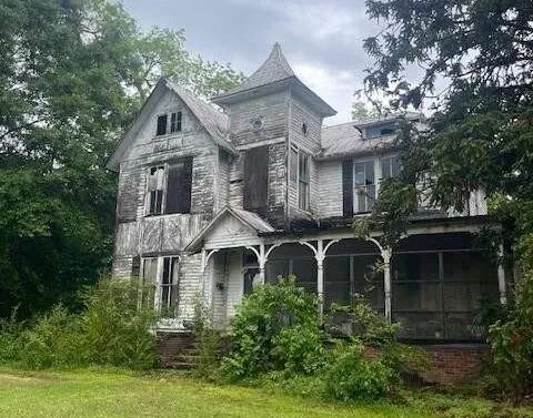 An abandoned two-story Victorian-style house with peeling white paint, broken and missing windows, and an overgrown front yard. A central tower with a steep roof rises above a covered porch, while dense trees surround the weathered home.