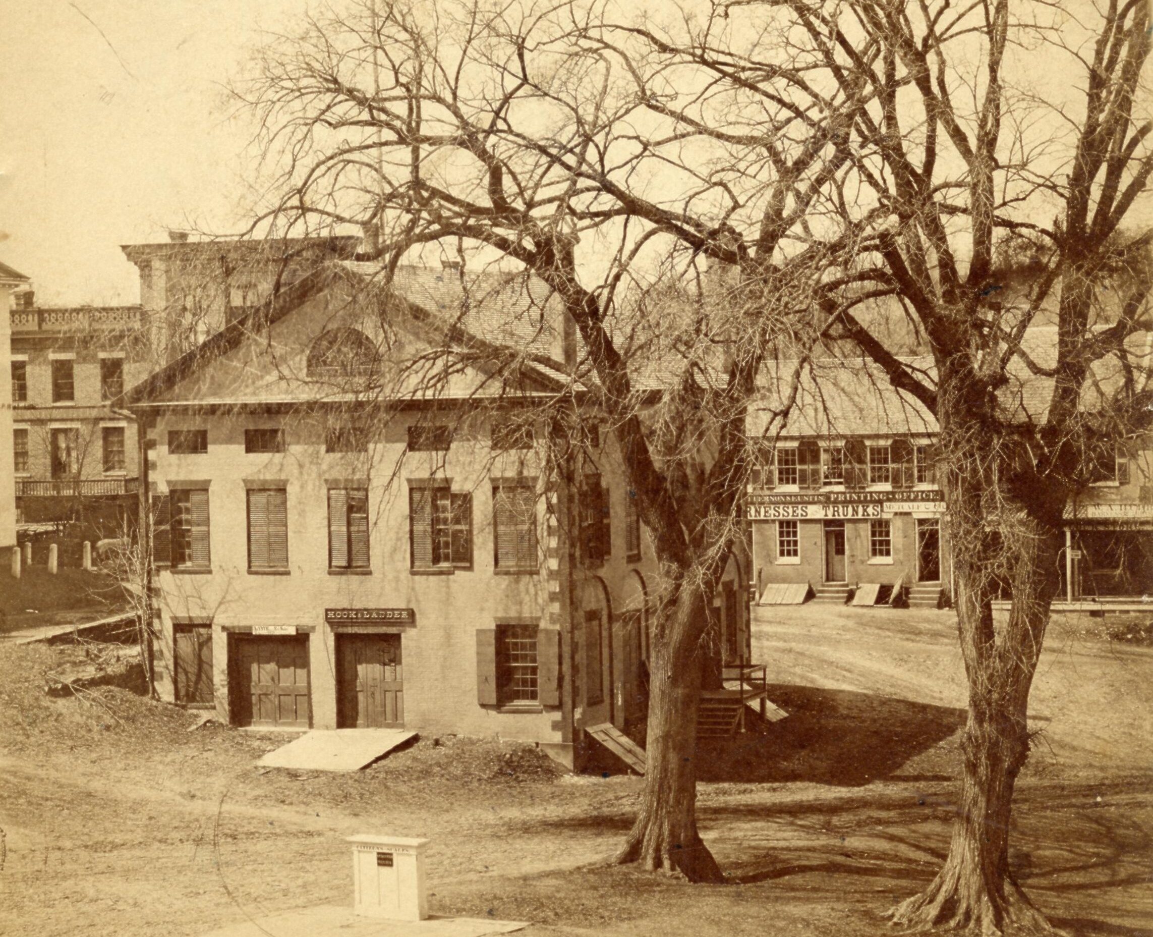 Black-and-white photograph of old town hall with stores lined behind it. The building has a slanted roof and wide, stable-like doors, one with a ramp infront of it. The sign over the front door reads “Hook & Ladder.”