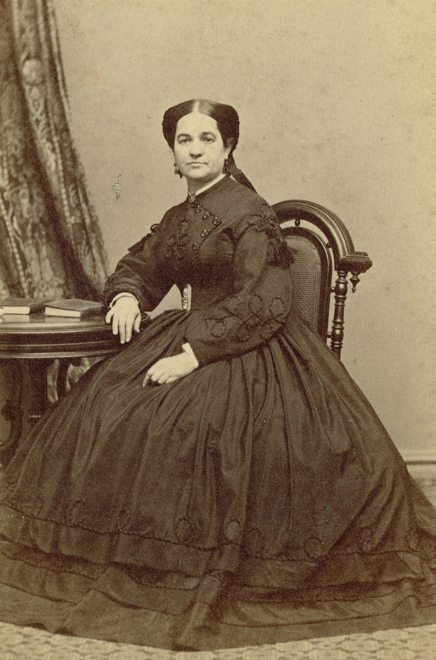 Black-and-white photographic portrait of a woman sitting at a small table with books on it. She is wearing a large black dress and has her hair in an intricate updo. One of her arms rests on the table and the other on her lap.