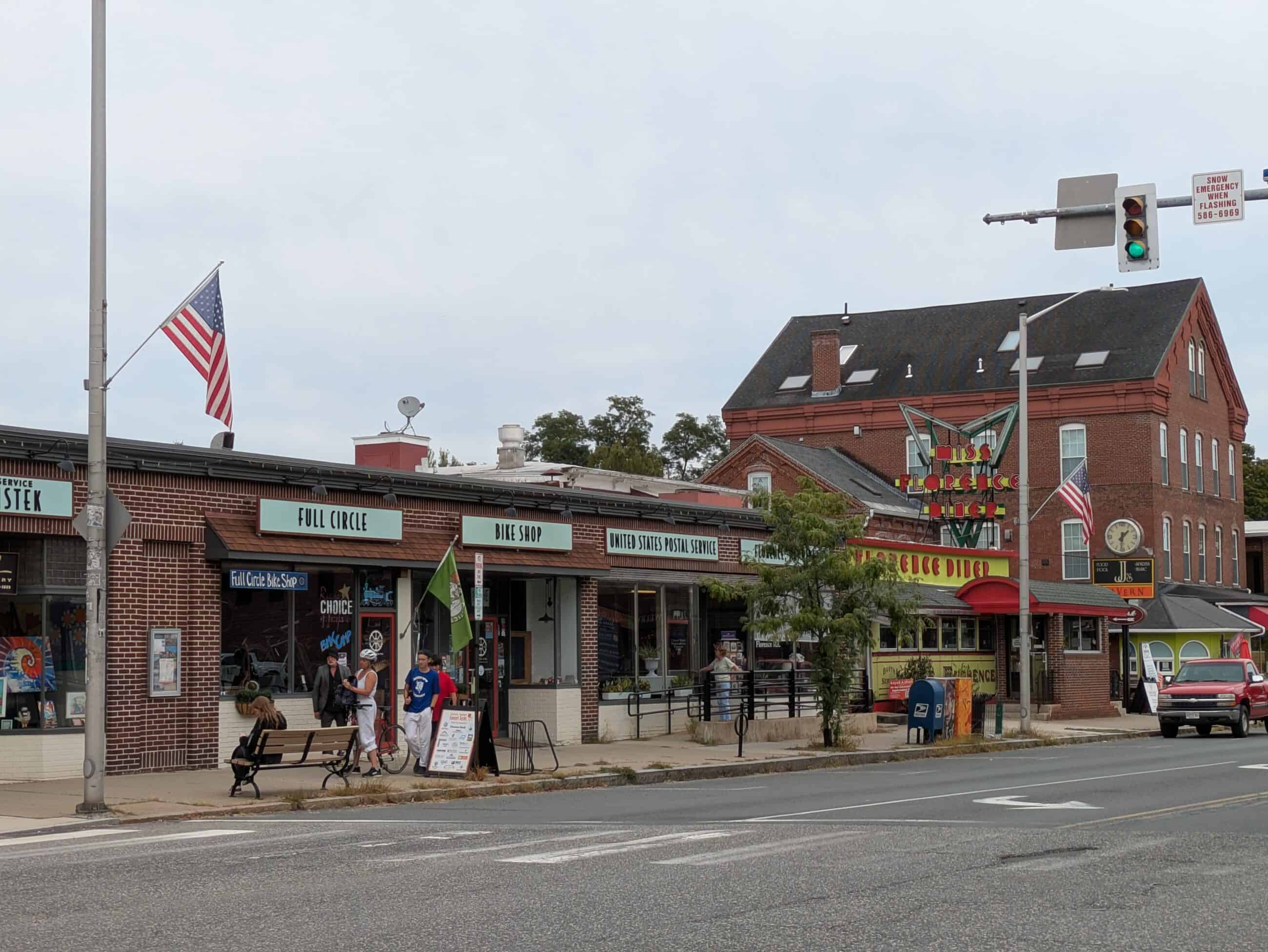 A modern photograph of Goodwin Block. Store fronts including Miss Florence Diner, Full Circle, "Bike Shop," and United States Postal Service are in view. All store signs are light blue and in same style except for Florence Diner. People walking and biking infront of the shops.