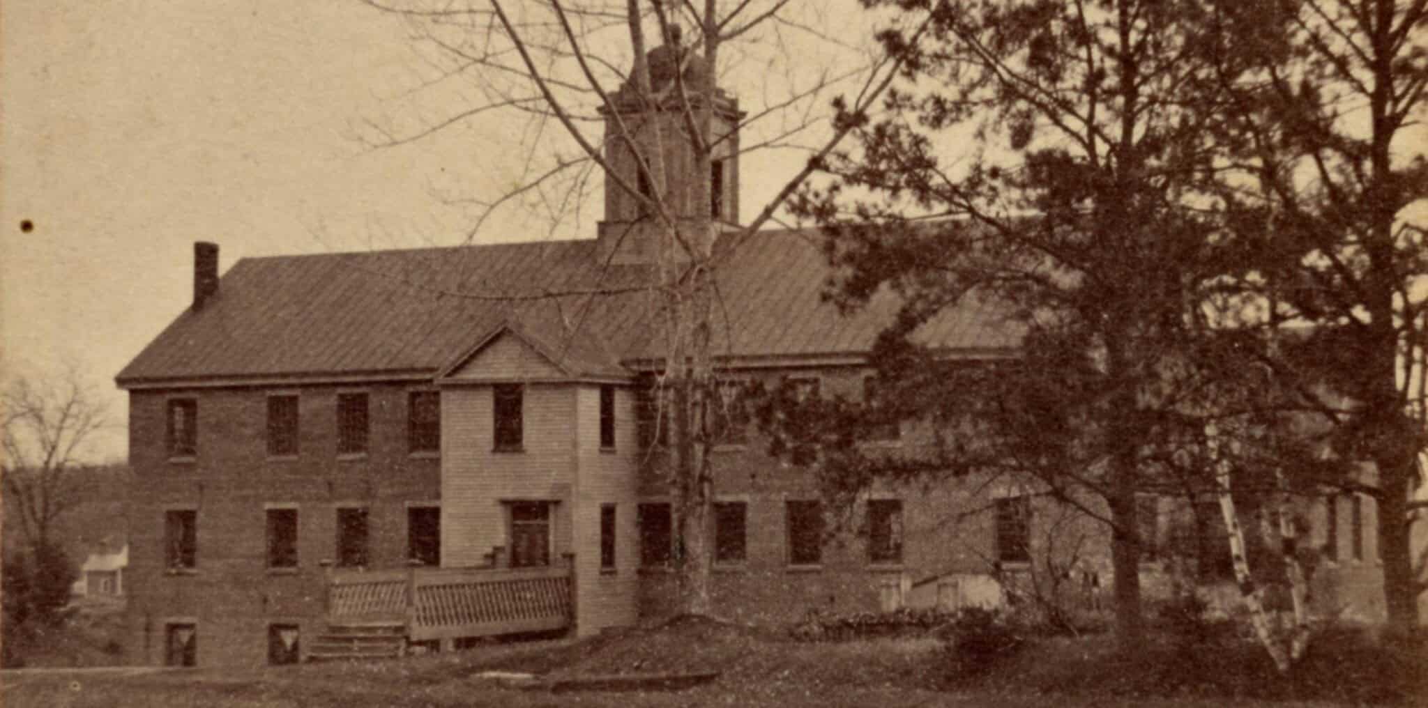 The photograph shows a structure that features a symmetrical layout with multiple evenly spaced windows across two main stories and a basement level. A prominent cupola or bell tower sits atop the gables in the center of the roof line. A wooden ramp and stairs lead up to an entrance on the left portion of the building's facade. Bare deciduous trees and evergreens partially obscure the front of the structure, which is set on a grassy hill.