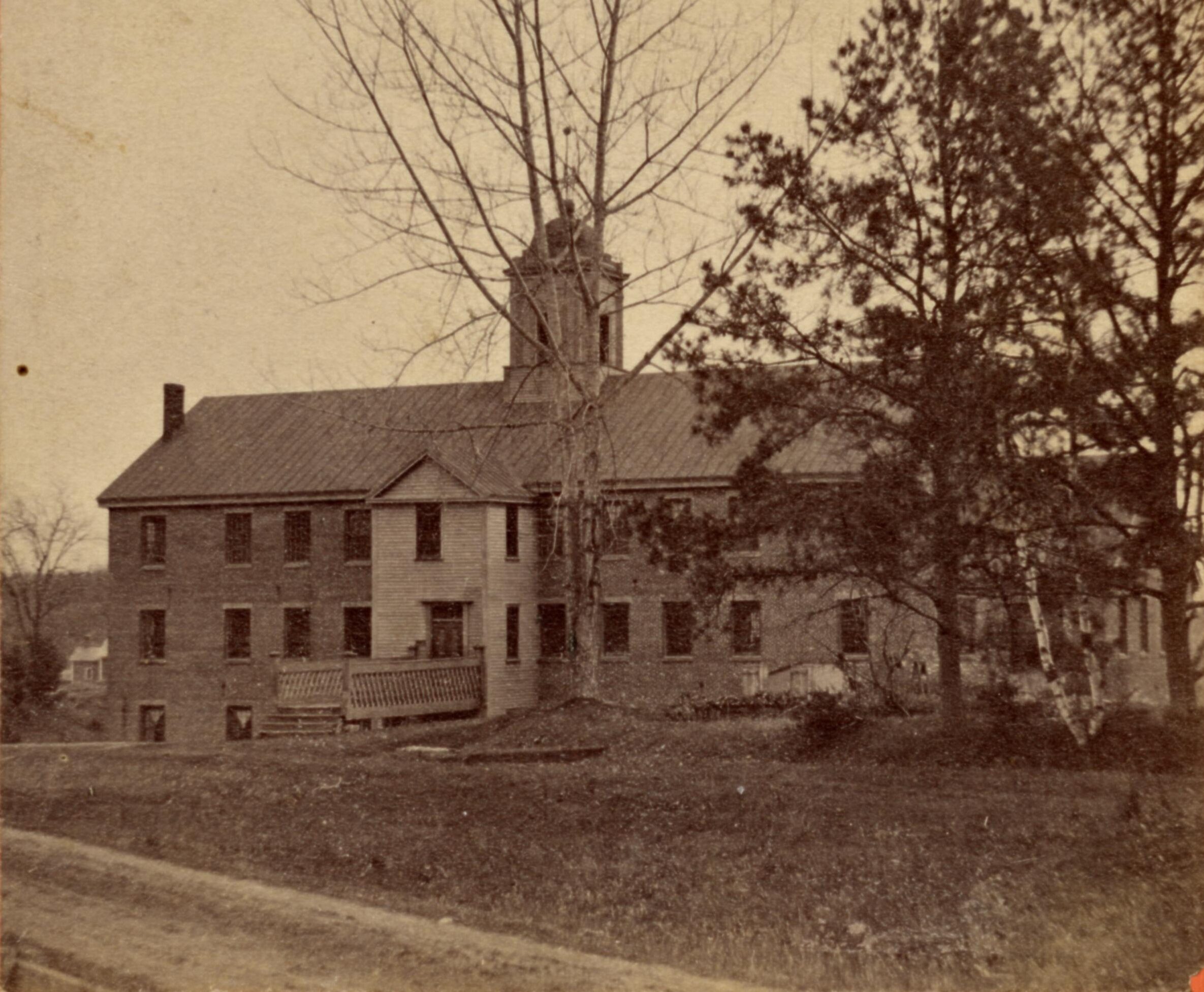 Photograph of the boardinghouse and silk factory of NAEI. The building appears to be brick and is rectangular with a slanted roof and a bell tower. Three trees stand in front of the building.