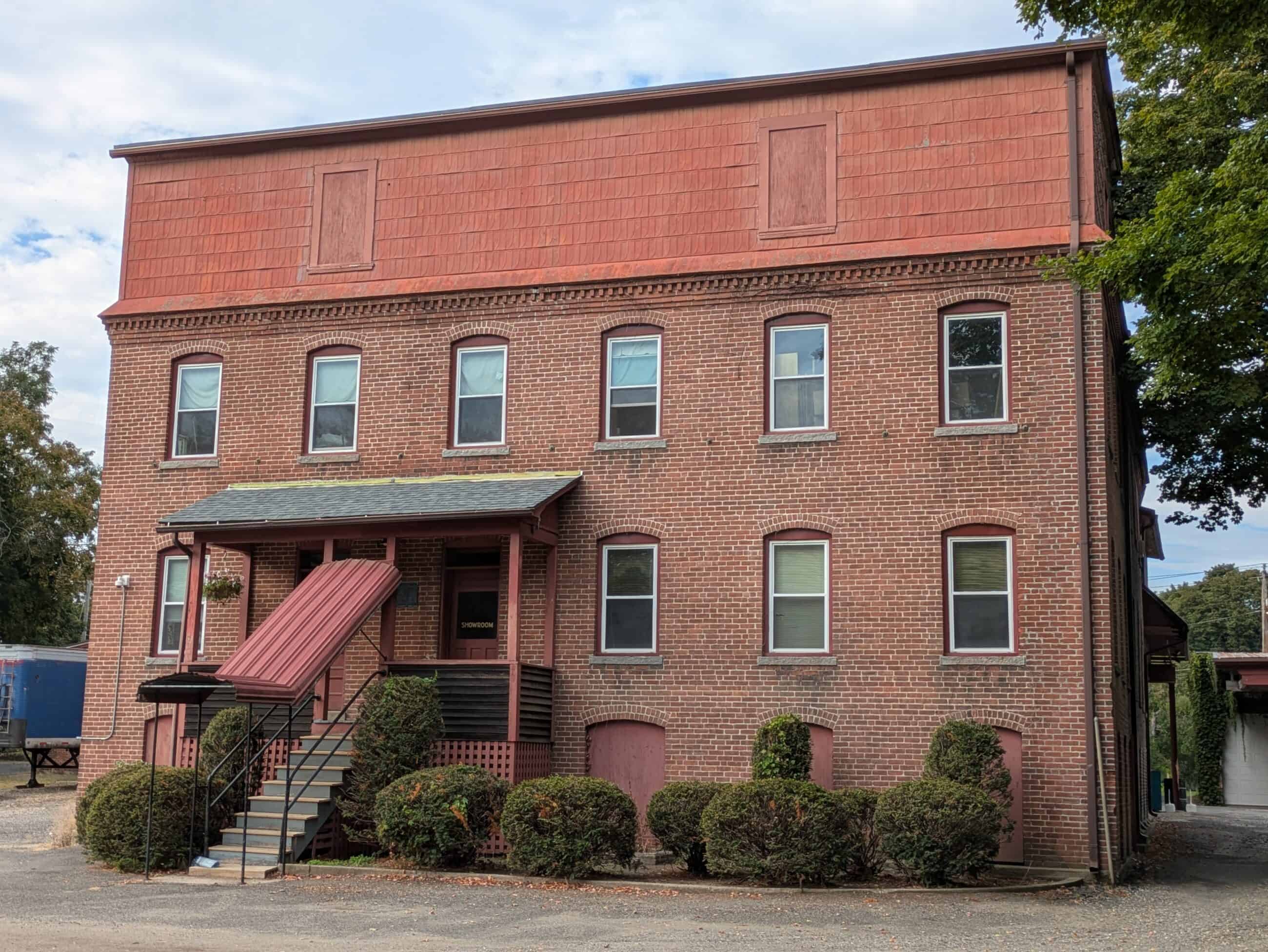 A photograph of the Florence Casket Company building. The building is old and red brick. There is a small porch infront. The front door has a sign that reads: "SHOWROOM."