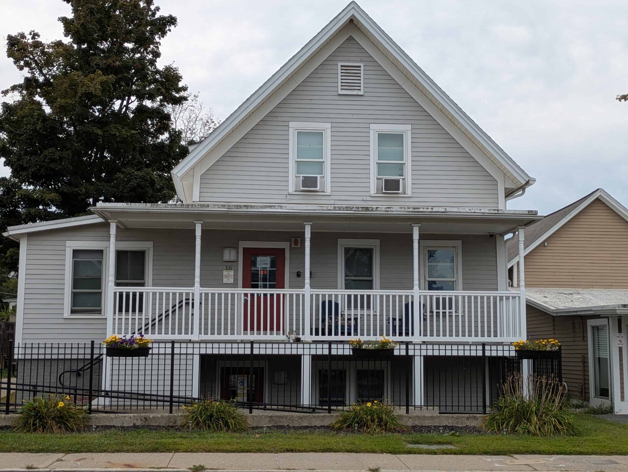 A photograph of the Asa Squires Building. The building is off white with a red door and with a slanted roof. There is a porch and a basement entrance visible in the photo. 