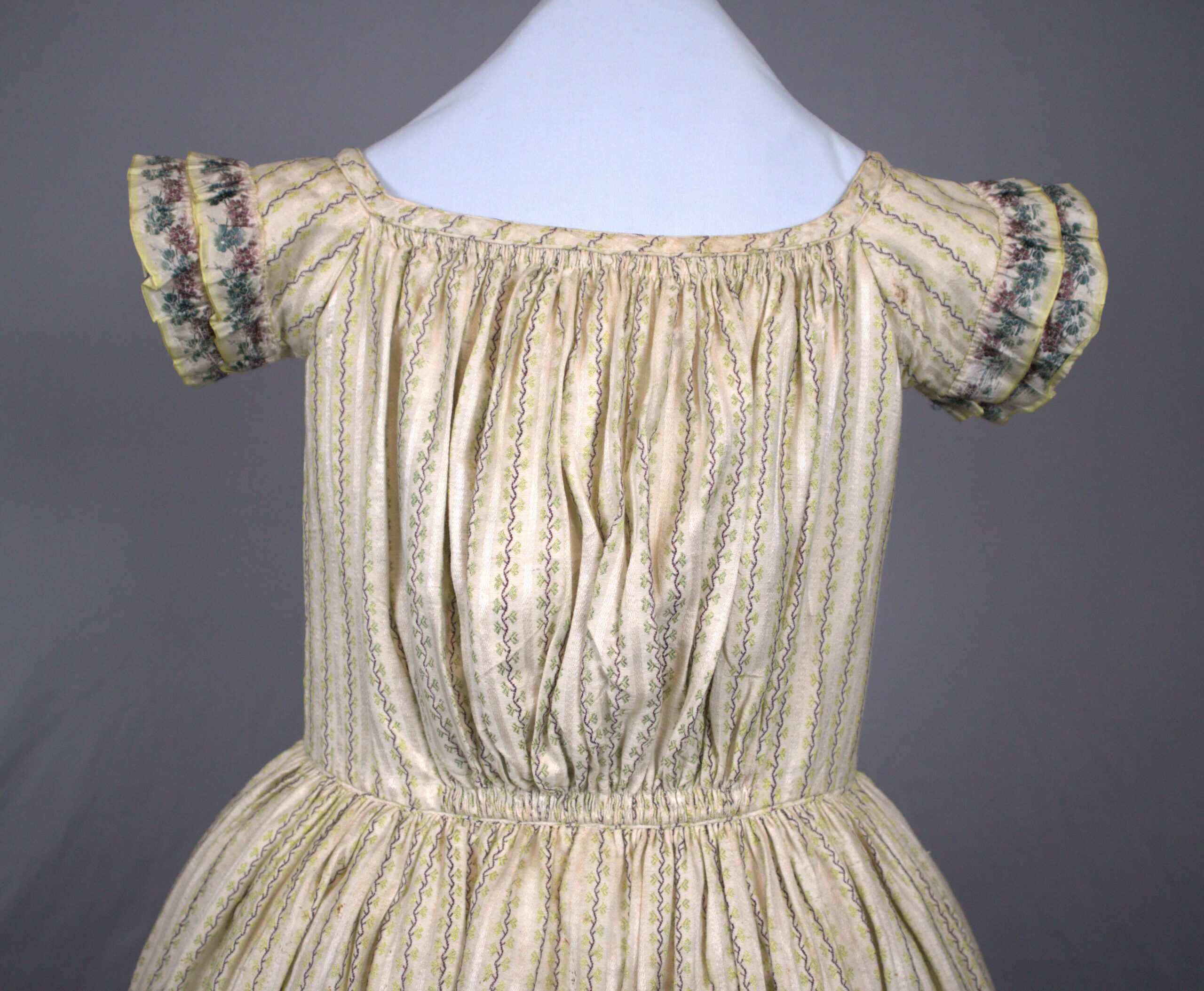 A close-up photograph of a cream-colored, 19th-century child's dress on a white mannequin torso against a solid grey background. The dress features a gathered, square neckline and short, ruffled sleeves with a decorative, floral-patterned trim in shades of green, pink, and yellow. The fabric is patterned with delicate, vertical stripes of tiny green and purple floral vines. The bodice is gathered tightly at both the neckline and the high waistband, creating soft folds down the center, while the skirt is gathered full at the waist.
