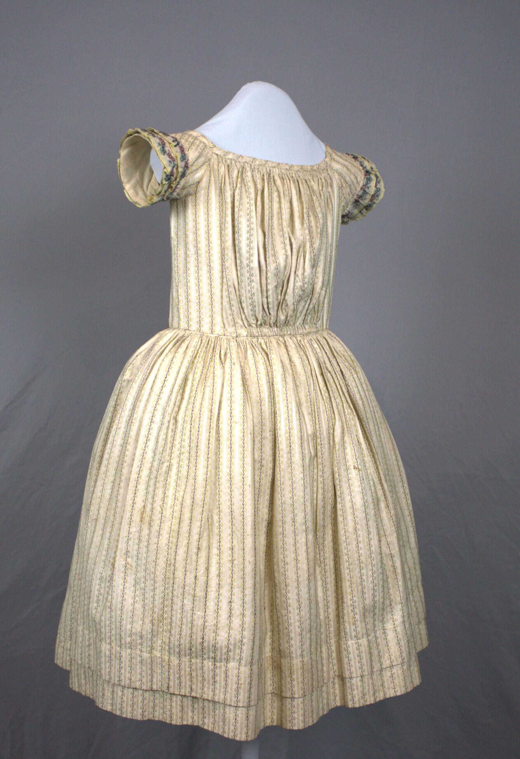 Young girl's dress made of cotton woven with a pattern of silk stripes of alternating purple and chartreuse vines on a white background. The short sleeves are bordered with two rows of ruffles made of silk ribbon woven with a floral design in purple and dark turquoise. The skirt is gathered very full all around the waist seam.