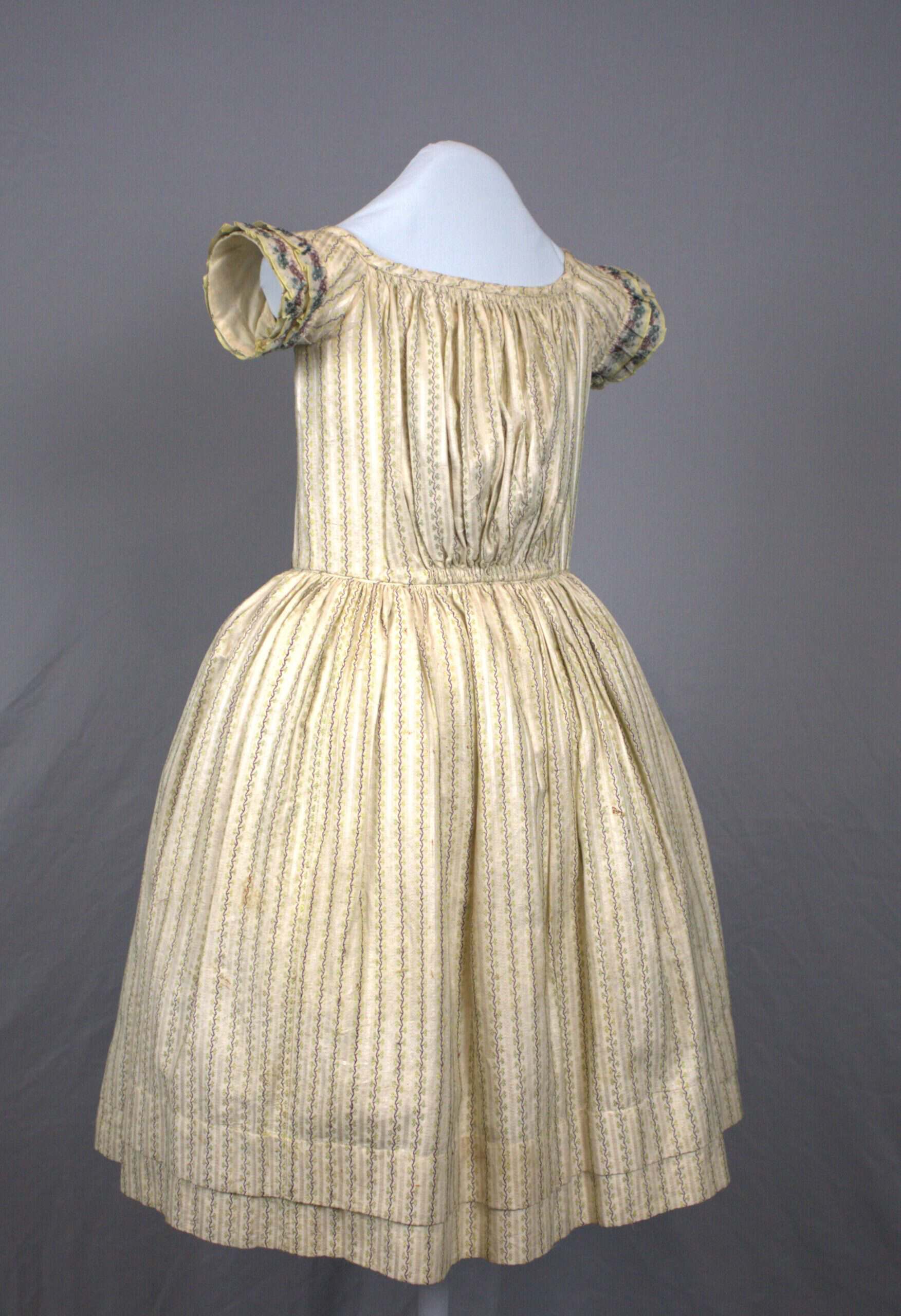 Young girl's dress made of cotton woven with a pattern of silk stripes of alternating purple and chartreuse vines on a white background. The short sleeves are bordered with two rows of ruffles made of silk ribbon woven with a floral design in purple and dark turquoise. The skirt is gathered very full all around the waist seam.
