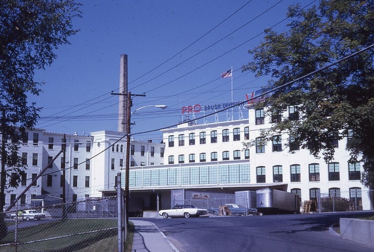 A photograph of the Pro Brush Division factory complex, a large white multi-story industrial building featuring a tall, slender chimney stack and an American flag flying on its roof under a clear blue sky. On the roof, red and blue neon signs spell out "PRO BRUSH DIVISION." The building has numerous rectangular windows and a low, wide entrance with two semi-trucks parked in front. A white classic car is parked near the entrance, and power lines crisscross the sky in front of the building. In the foreground, a paved street curves toward the right, with a chain-link fence on the left bordering a green lawn.