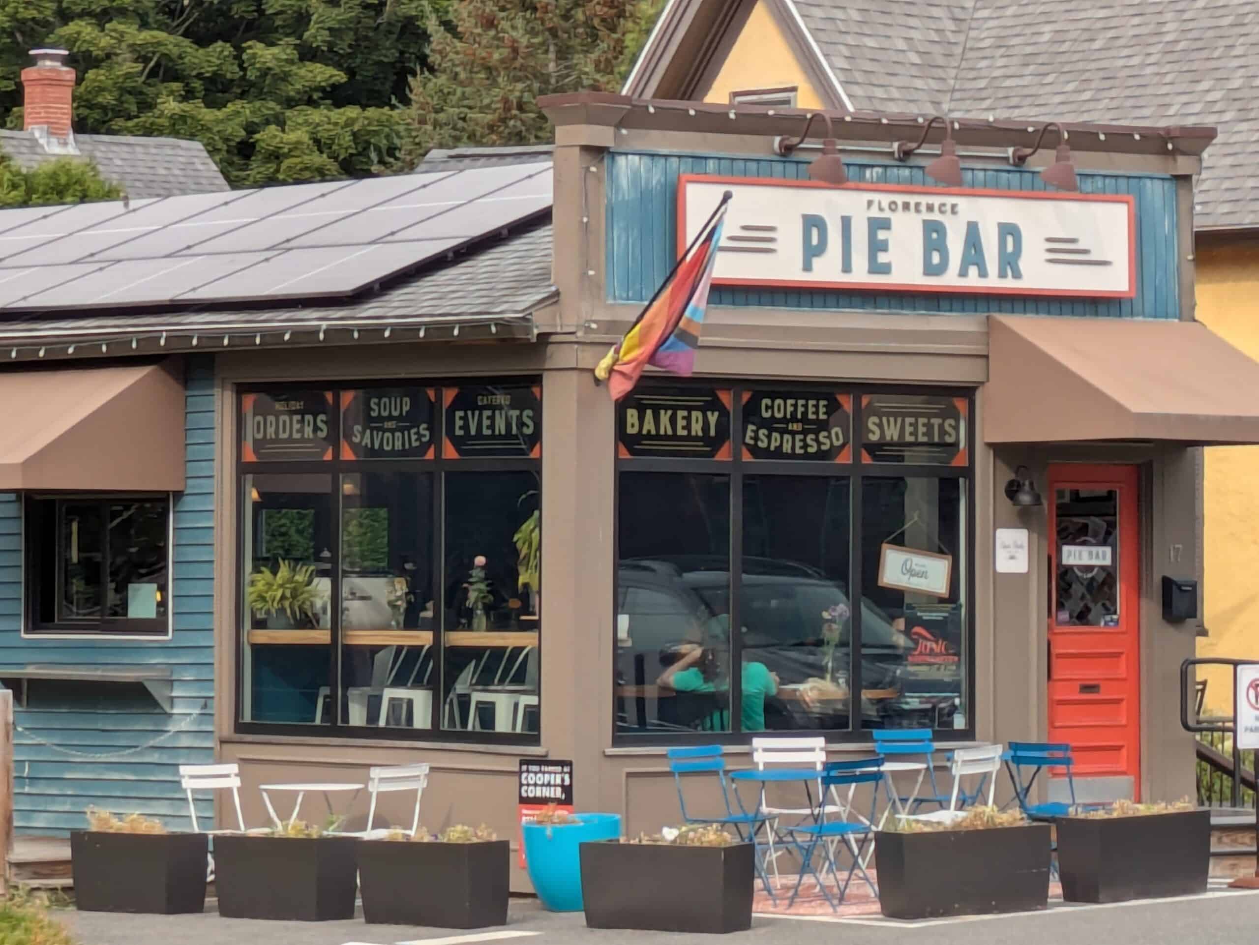 A photograph showing the exterior of the Florence Pie Bar, a single-story bakery and cafe housed in a rustic, wood-paneled building. The building’s facade features a mix of weathered blue siding on the left and a warm, tan-painted storefront on the right. A large, prominent rectangular sign reading "FLORENCE PIE BAR" in bold, white capital letters against a blue background is mounted above the main windows, flanked by three stylized horizontal lines on either side. Below this sign, six large, dark-framed glass window panes showcase the cafe's offerings with smaller, decorative signs reading "HOLIDAY ORDERS," "SOUP AND SAVORIES," "CATERED EVENTS," "BAKERY," "COFFEE AND ESPRESSO," and "SWEETS." A rainbow flag hangs at an angle near the center of the roofline. To the right, a bright orange-red door with window panes serves as the entrance, next to a black handrail on a small set of steps. In front of the building, an outdoor seating area is arranged on the asphalt, featuring several white and blue folding metal chairs and small tables. A row of large, dark rectangular planter boxes filled with low-growing plants lines the front of the seating area.