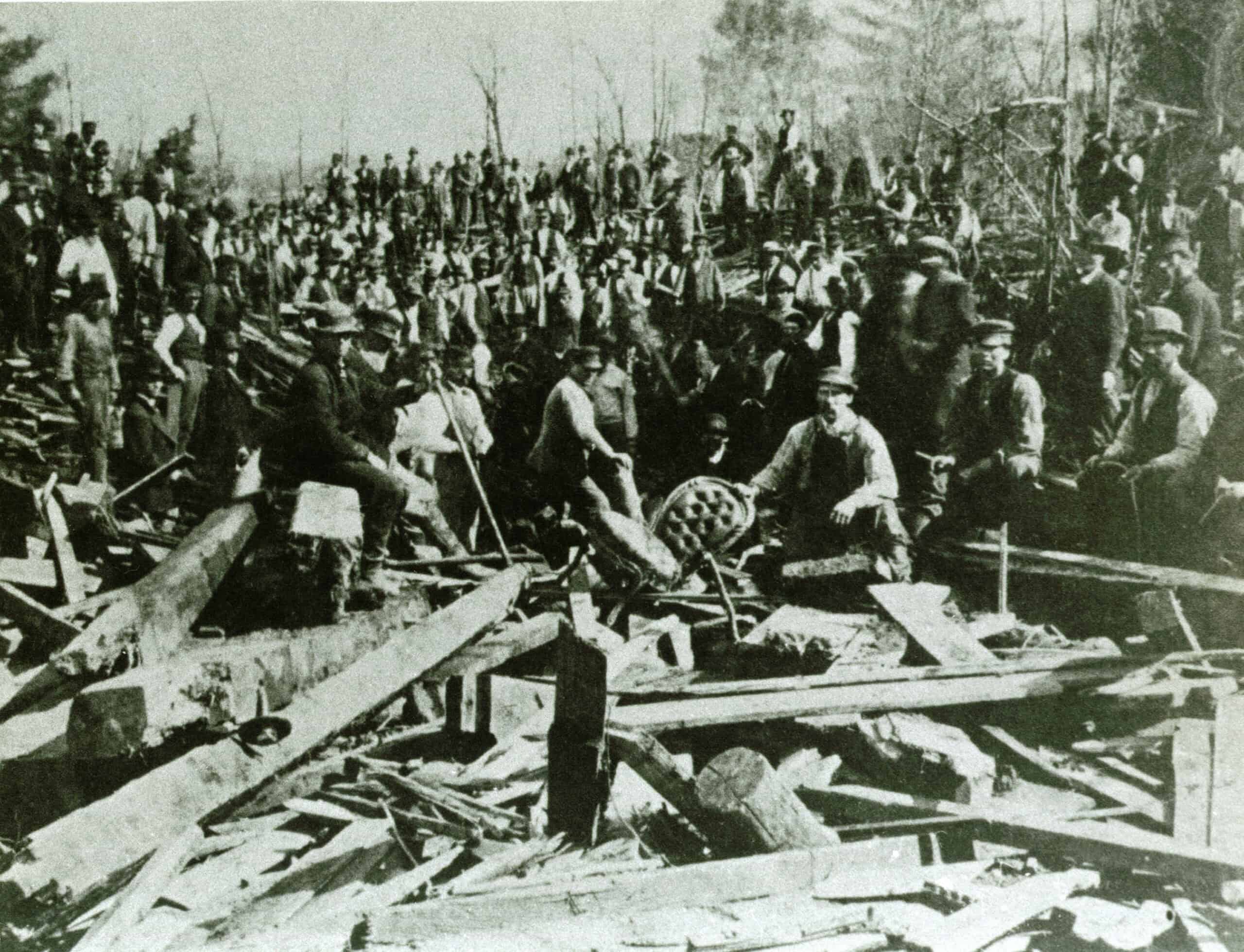 A black and white photograph shows a large crowd of men gathered around a massive field of debris, composed of shattered wooden planks, beams, and logs. In the foreground, several men sit or stand directly amidst the wreckage; one man near the center holds a rounded object, possibly a piece of machinery or household item salvaged from the destruction. Behind them, dozens of workers and onlookers stand in densely packed rows on a hillside, overlooking the devastation. 