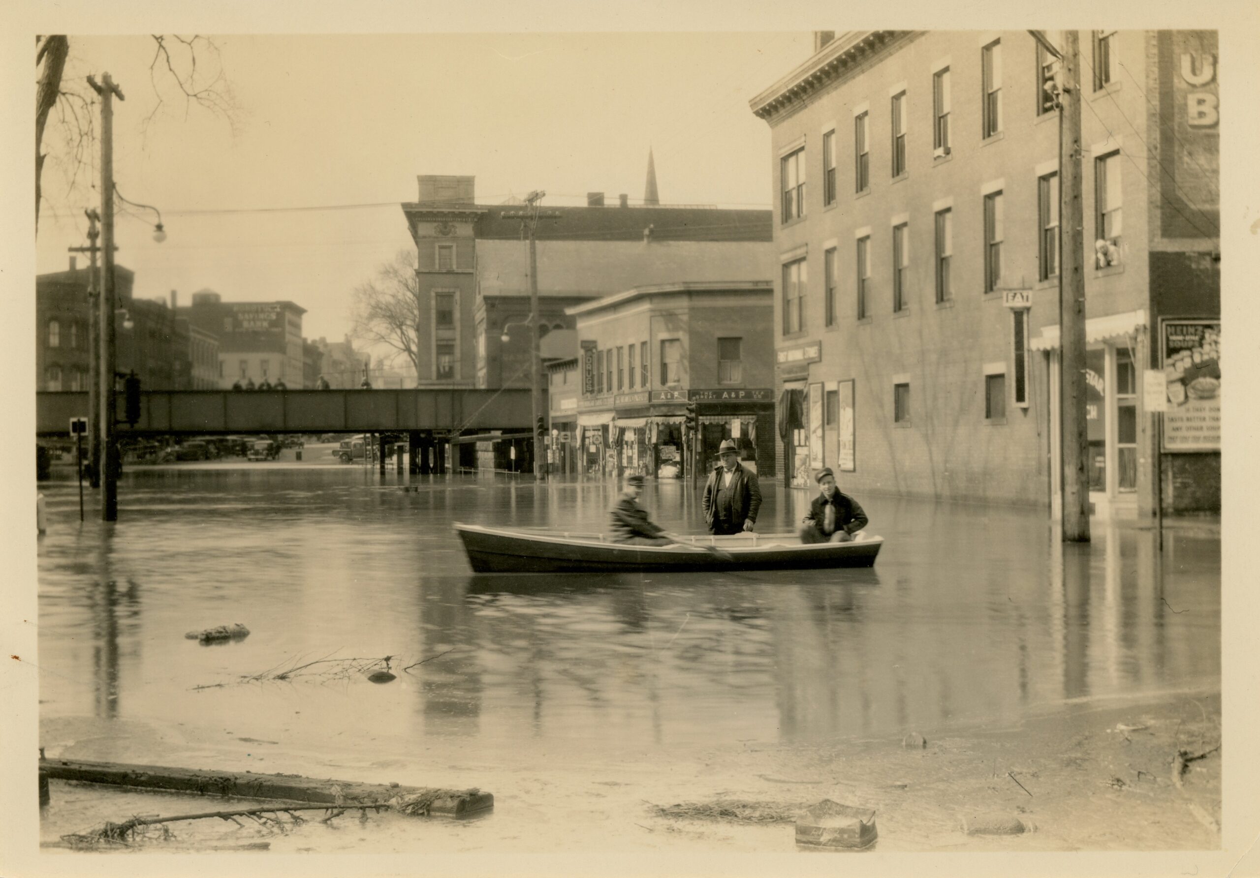 3 men on a small boat in the water in the middle of Main Street with the railroad bridge in the background