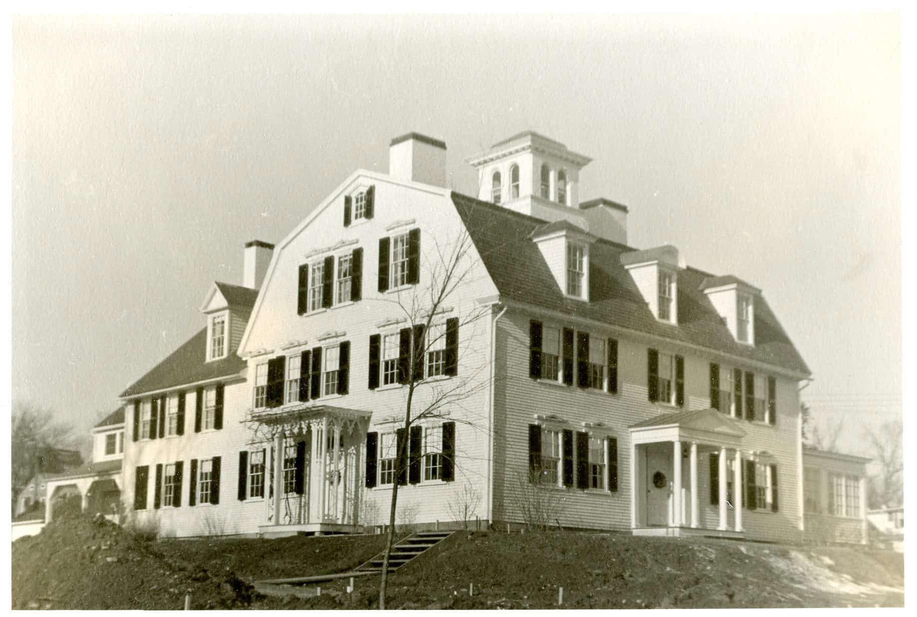 Photograph of the exterior front of the Manse. The photograph is black-and-white and shows the Manse house, which is white with dark shutters. The roof is brick and has three protruding windows. Above the front door is a marquee with 4 columns in a Romansesque style. A marquee covering the side door can also be seen, in a more decorative style. A small watchtower peaks through the roof of the house.