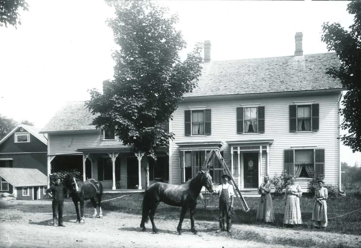 Black-and-white photograph showing a farm family posed outdoors in front of a large, two-story farmhouse. The house is a large white clapboard home with dark-shuttered windows, two brick chimneys, and an ornamental front porch entry. An attached lower wing extending to the left features a covered porch with supportive pillars. A dark barn and a small shed are in the background, but mostly cut off. On the left, standing in front of the attachment of the house, a young man stands beside a dark horse with white lower legs and hoofs. Toward the center of the photograph, in front of the main house building, a man wearing overalls holds the lead rope of a second horse standing broadside. On the right of him, three women dressed in long, floor-length patterned dresses and aprons stand side-by-side near the front lawn. A wooden A-frame ladder rests against the side of the house behind the central horse and man.