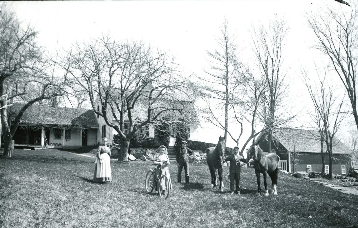 Black-and-white photograph of a family with two horses, in front of their house. A little girl has her bicycle.