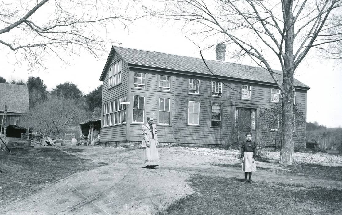 A black and white photograph of a two-story wooden clapboard house situated on a gentle slope, with two people standing outside on a dirt pathway. The house features a gabled roof, a brick chimney on the right side, and numerous multi-pane sash windows across its facade. On the left side of the house, an older woman stands facing forward, dressed in a long, light-colored Victorian-style dress with a matching jacket. To the right, closer to a large bare tree in the foreground, a young girl stands facing the camera, wearing a dark dress with a light-colored apron. In the background to the left, a portion of a barn and another small outbuilding are visible amidst sparse trees.
