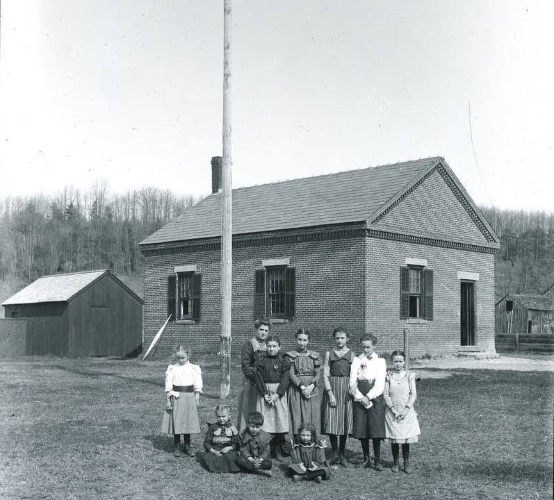 A black and white photograph captures a group of ten children standing and sitting on a grassy lawn in front of a small, one-story brick schoolhouse. The children are dressed in historic attire from the late 19th or early 20th century. Six older girls stand in a line across the center, wearing long dresses or skirts paired with white blouses. Four younger children are positioned in the foreground; two girls stand on the outer flanks, while two younger boys sit cross-legged on the grass in the center.The rectangular schoolhouse features a gabled shingle roof with an ornamental brick dental molding along the roofline, a chimney at the rear left, and dark wooden shutters on its multi-paned windows. A very tall, slender wooden flagpole stands prominently in the yard to the left of the building's front facade. The surrounding background consists of a clear sky, a line of leafless trees on a distant ridge, a small wooden outbuilding on the left, and a partial view of another rustic wooden building on the far right.