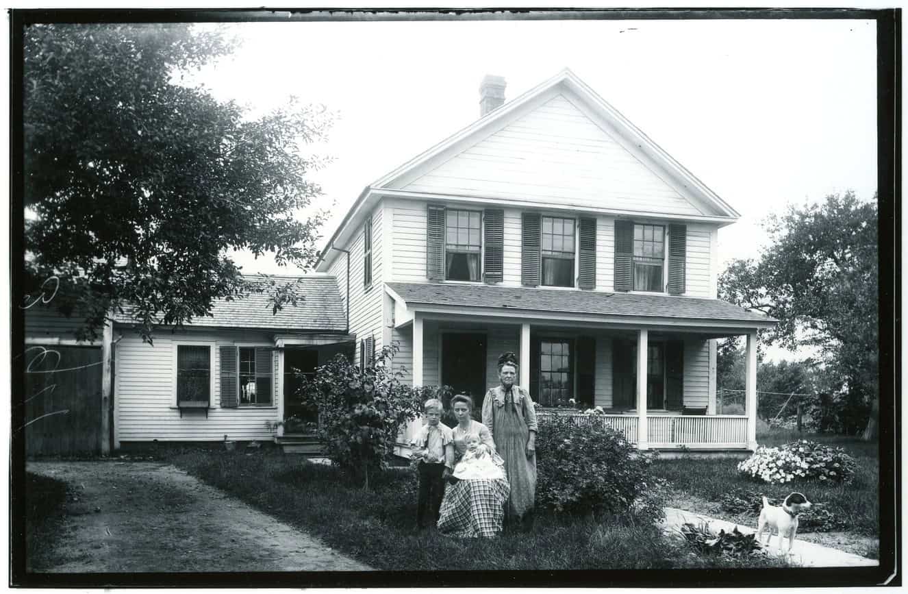black and white photograph of a family in front of a clapboard house with a front porch and garden. A woman is seated in the foreground with a baby on her lap, a young boy stands on one side of her, and an older woman stands on her other side. A terrier dog stands on the walkway
