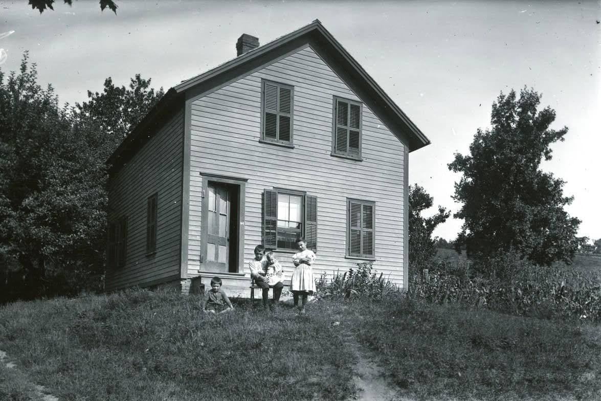 A black and white photograph shows a family of four posing in front of a modest, two-story wooden clapboard house situated on a grassy hill. The house features a steeply pitched gabled roof with a central chimney and shuttered windows on both levels. To the left of the house, an open wooden door reveals a dark entryway. In front of the house, a woman in a light-colored dress stands holding an infant, while a man sits beside her on a small bench, holding a young child on his lap. Another young boy sits on the grass just below them, facing the camera. The property is surrounded by dense trees on the left and a cultivated field or tall vegetation on the right.
