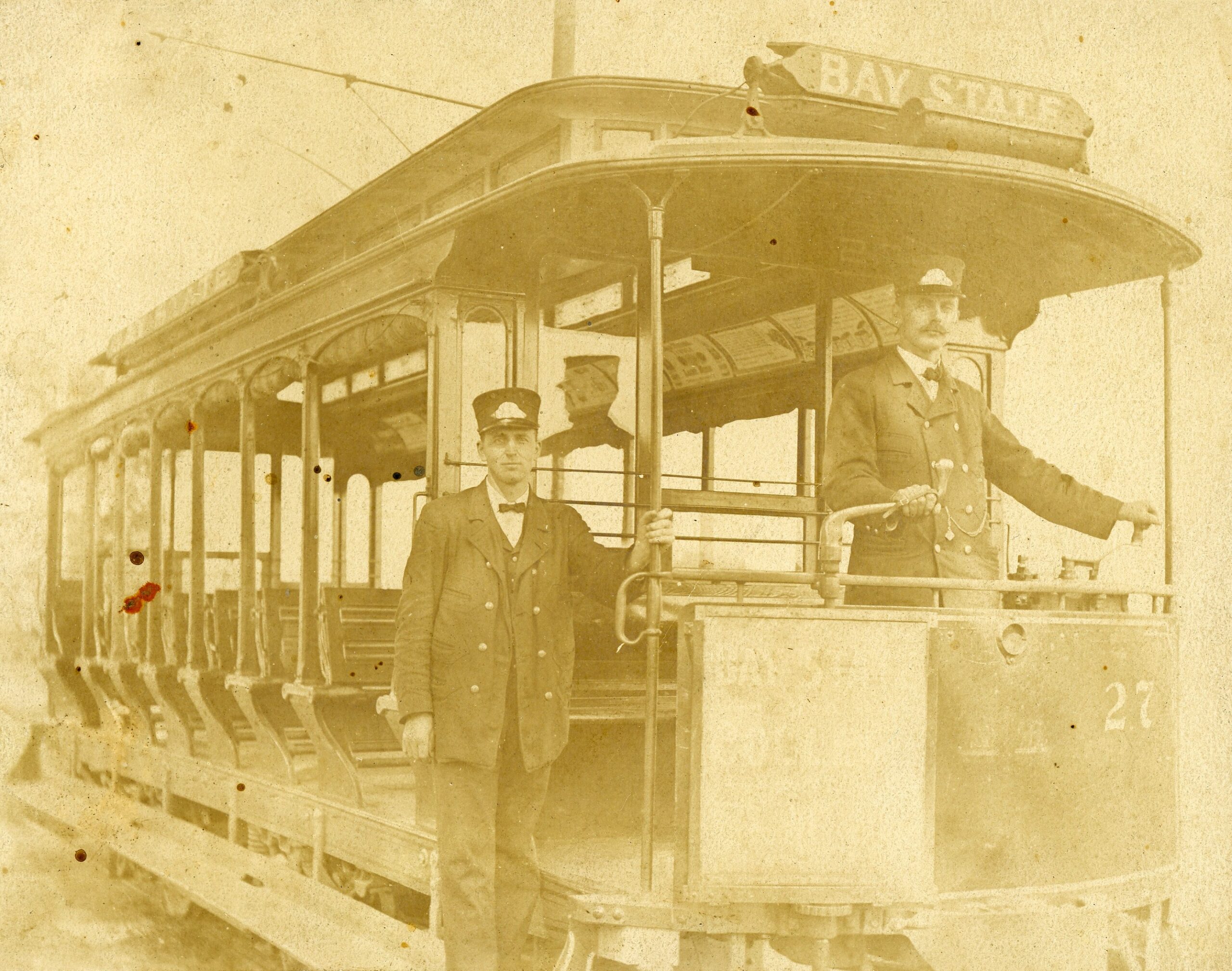 An extremely yellowed black-and-white photograph of an open-air streetcar numbered 27, branded with a "BAY STATE" sign on its roof. Two uniformed transit workers pose with the vehicle; one conductor stands on the ground alongside the open passenger rows, while the motorman stands at the front controls on the platform. The open-sided trolley features rows of wooden bench seats under a curved roof supported by slender pillars, with overhead electric wires visible above.