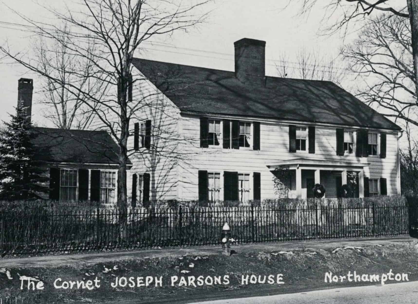A black-and-white photograph captures the front view of the the Parsons House, taken from across the street. The main structure is a two-story Colonial-style home with a side-gabled roof, light-colored horizontal siding, and a large brick chimney emerging from the center of the roofline. A small, covered front porch with simple white columns shelters the central entrance, adorned with two dark holiday wreaths. To the far left, a smaller, one-story gabled wing attaches to the main house, featuring its own end chimney and shuttered windows. In the foreground, a dark wrought-iron fence separates the property from a dirt roadside bank, where a single fire hydrant stands. Bare, leafless winter trees flank both sides of the house. Handwritten text in white cursive script at the bottom left reads "The Cornet JOSEPH PARSONS HOUSE," while "Northampton" is written at the bottom right.
