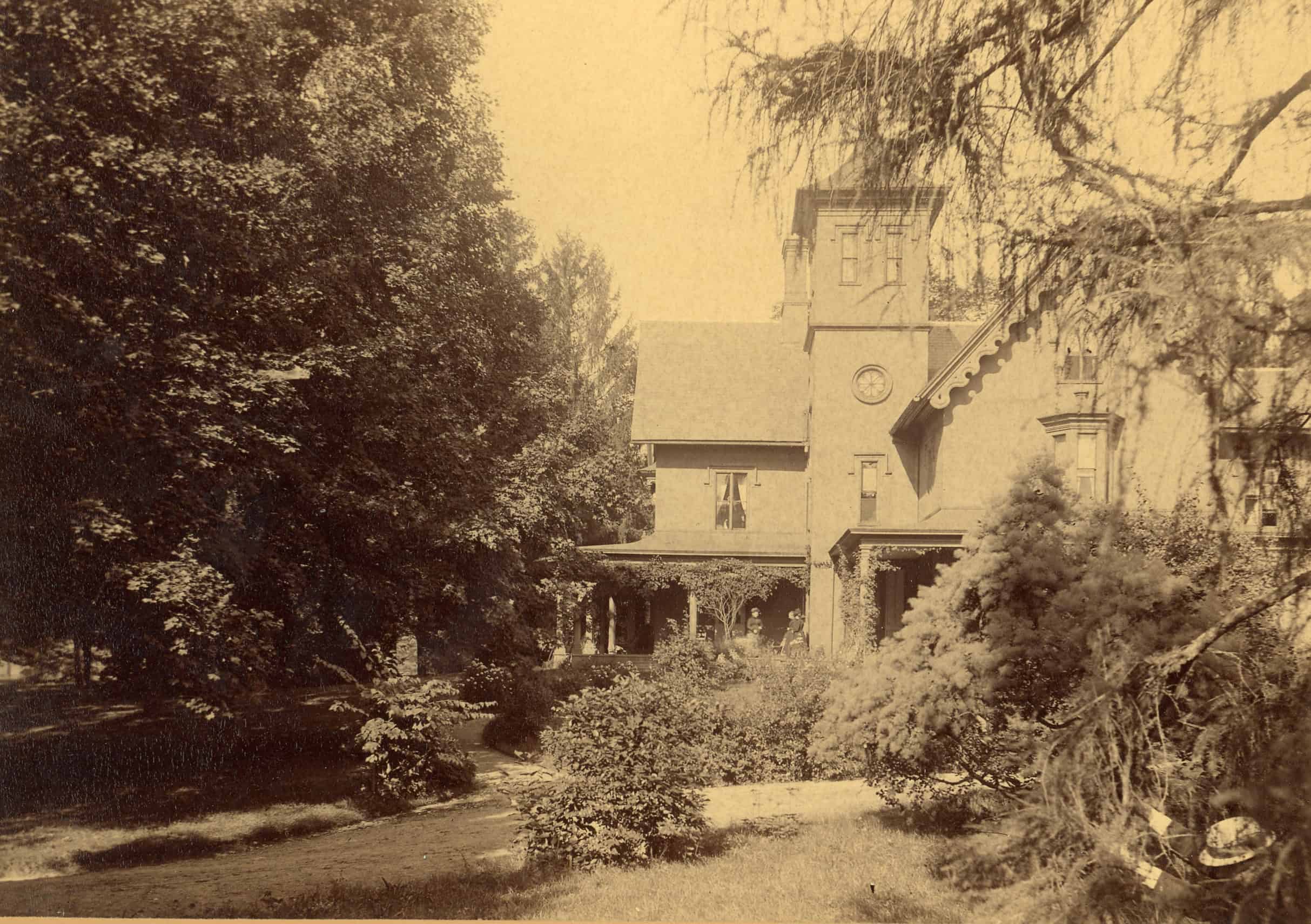 An old, yellowed photograph of a large, two-story Gothic Revival estate nestled behind lush, overgrown trees and shrubbery. The house features a prominent central square tower with windows at the top, a steeply pitched roof, decorative bargeboards along the gables, and a wide covered porch stretching across the front. A dirt path curves through the dense, leafy yard in the foreground under the shade of mature trees.