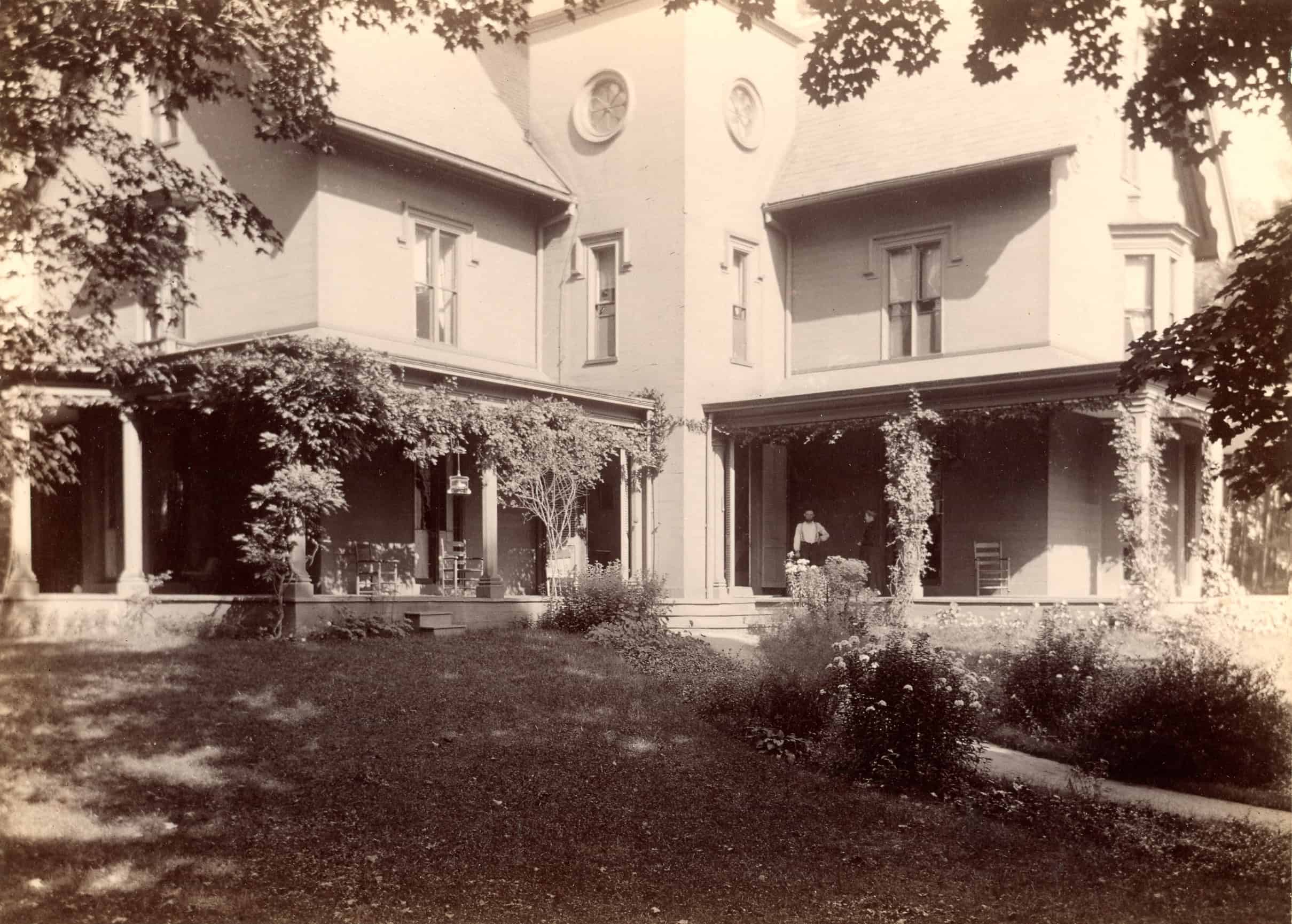 A black-and-white photograph captures the wide exterior of a grand, multi-story house viewed from a slightly low angle across a spacious, grassy lawn. The large residence features two primary wings meeting at a central, forward-projecting tower section. This central tower includes distinct round, portal-style windows near the roofline, while the main wings display tall, rectangular double-hung windows. A continuous, deep covered porch wraps across the front of both wings, supported by a series of classic, light-colored columns. Lush, climbing ivy and leafy vines heavily drape over the porch headers and columns. Two people are visible standing in the shaded porch area of the right wing. The house sits on a gently sloping grass lawn, with a curved dirt or gravel pathway visible on the right. Diverse flower beds, low shrubs, and ornamental plants are arranged near the porch foundation, while the upper corners of the frame are softly bordered by the overhanging leaves and branches of mature trees.