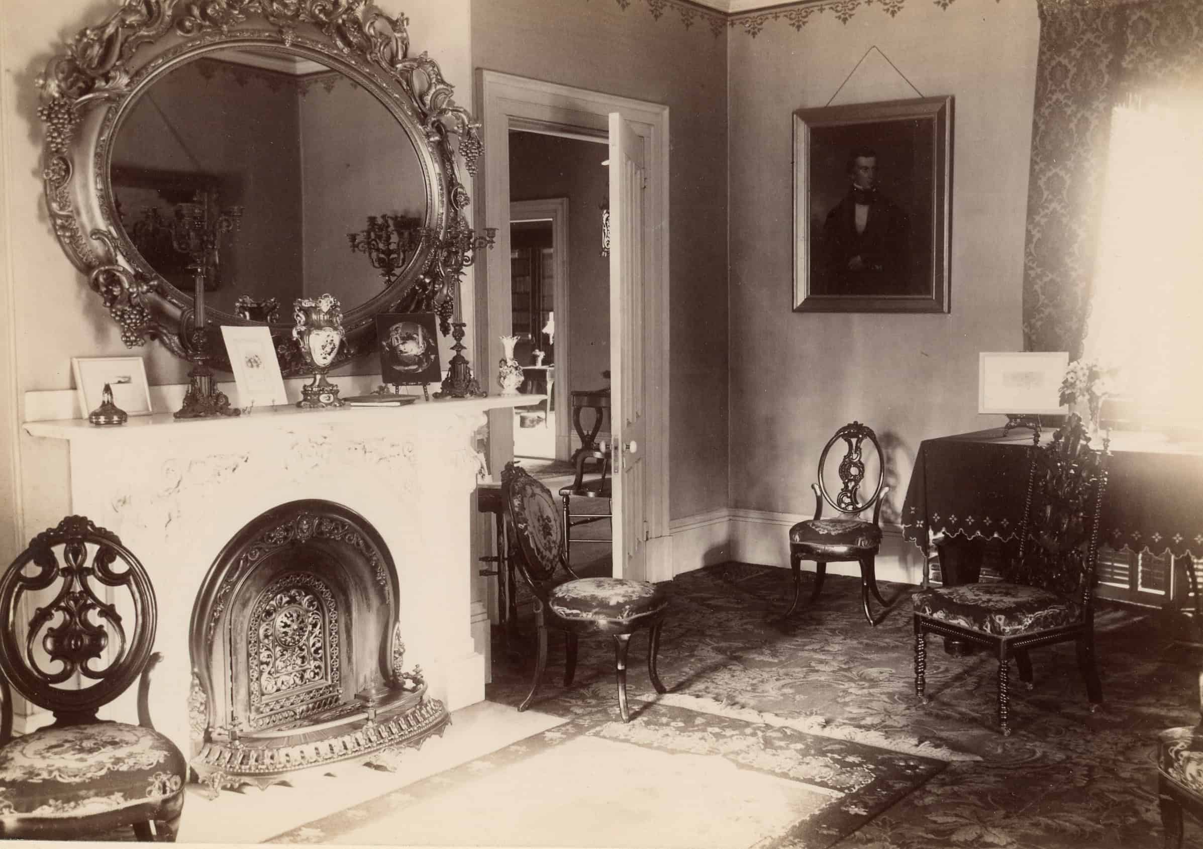 A black-and-white photograph capturing the interior of a Victorian-style parlor room. On the left side of the room stands a white fireplace with an intricate, dark metal grate insert. Mounted on the wall directly above the mantel is a massive, ornate oval mirror featuring a deeply carved, decorative frame. Several small framed pictures and decorative objects are meticulously arranged across the mantelpiece. To the right of the fireplace, an open doorway reveals a glimpse into an adjacent room. Scattered throughout the parlor are multiple high-backed wooden chairs, each featuring elaborate openwork carvings on the backrests and patterned fabric upholstered seats. The floor is covered with a large, patterned area rug. In the background on the right wall hangs a framed portrait of a man, positioned near a fabric-covered table placed beside a window draped with heavy curtains.