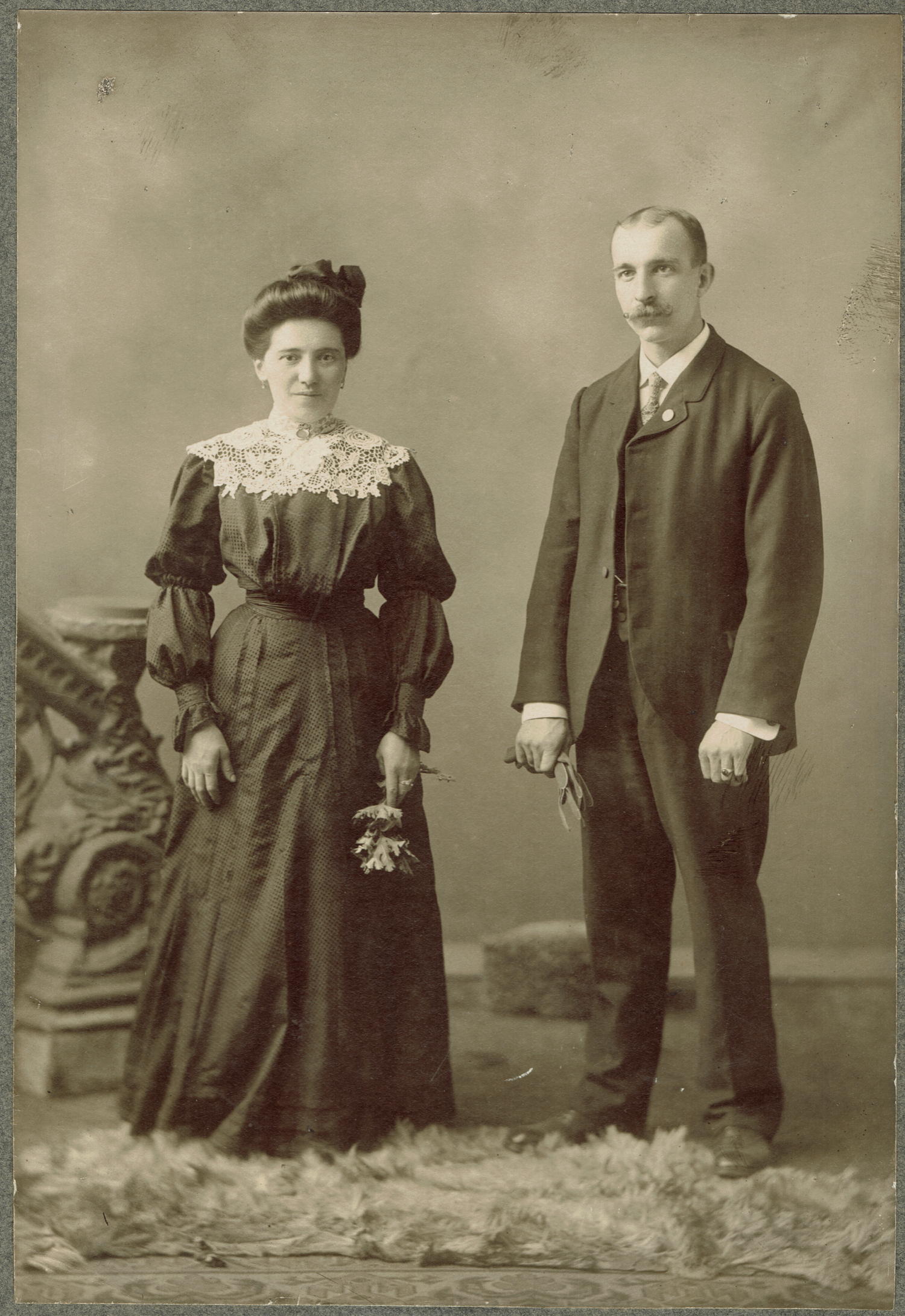 Black-and-white photographic portrait of a couple. Both the woman (left) and the man (right) stand on a small fur carpet. In the background the beginning of an ornate stair railing can be seen. The woman has her dark hair in an updo and is wearing a black dress cinched at the waist with puffy sleeves. From the shoulders to the top of the neck on the dress is white lace. The woman has her hands on her sides, with one holding a flower. The man is in a black suit and tie with a white undershirt and cuffs. He wears dress pants and black shoes. He has a prominent mustache and thinning hair. He is holding something in one of his hands.