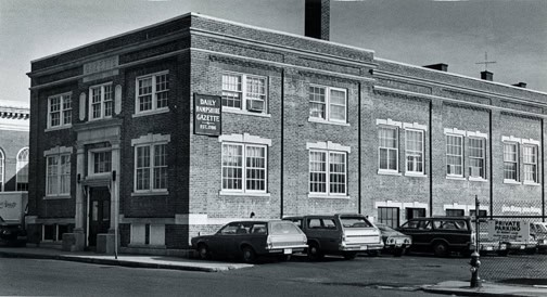 Black-and-white photograph of
the Hampshire Gazette building at 16 Armory Street, Northampton. The main part of the building is brick and square-shaped, with an extended brick addition part less wide and longer. A sign on the side of the building reads: "DAILY HAMPSHIRE GAZETTE EST. 1786" and "GAZETTE" is engraved into the top of the front exterior of the building. Cars are lined in the parking lot on the property.