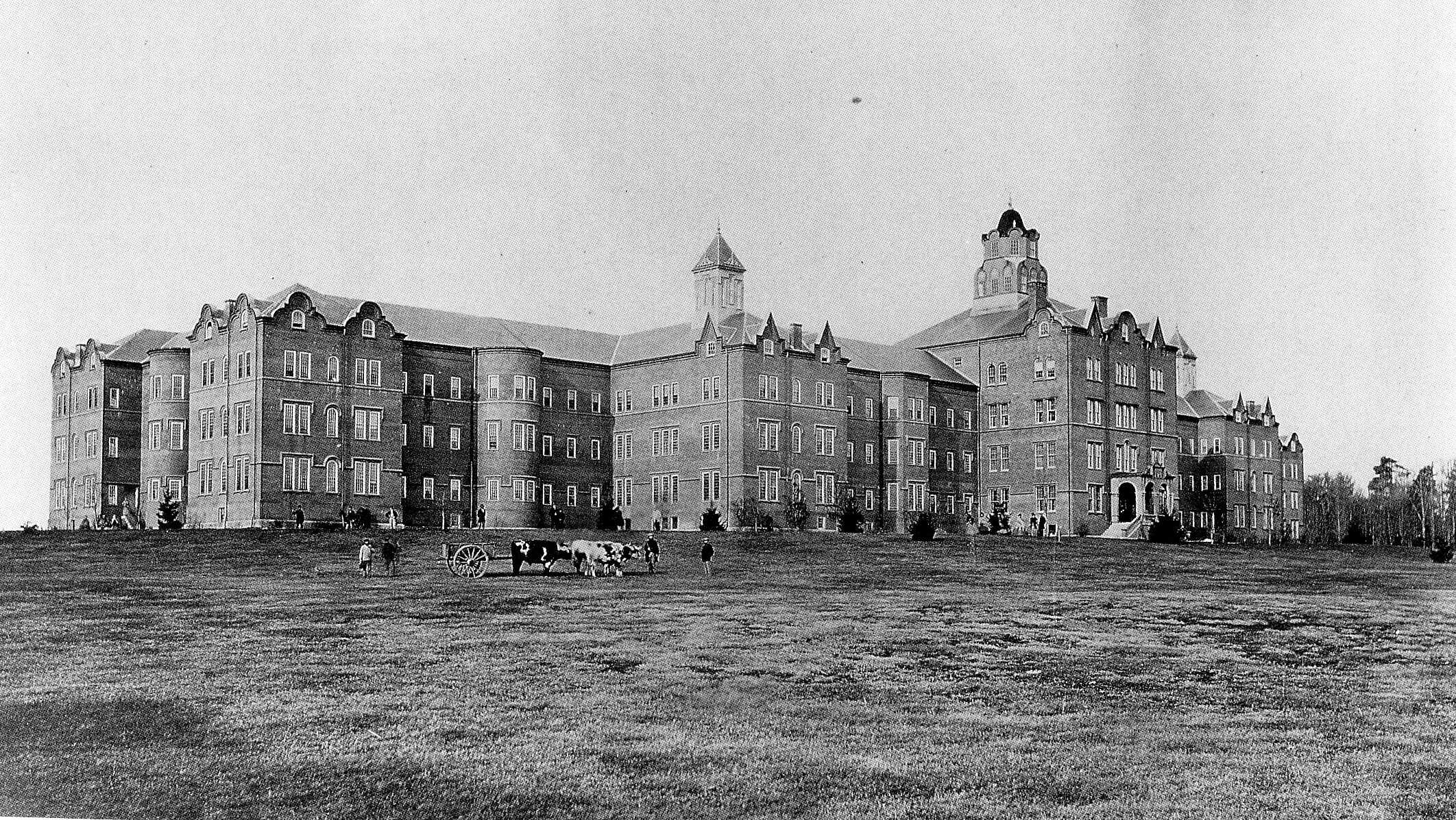 Black-and-white photograph of the exterior of Northampton State Hospital. The building is very large and appears to be made of brick. In front of the building live stock and carriages can be seen. The property is surrounded by a lot of land.