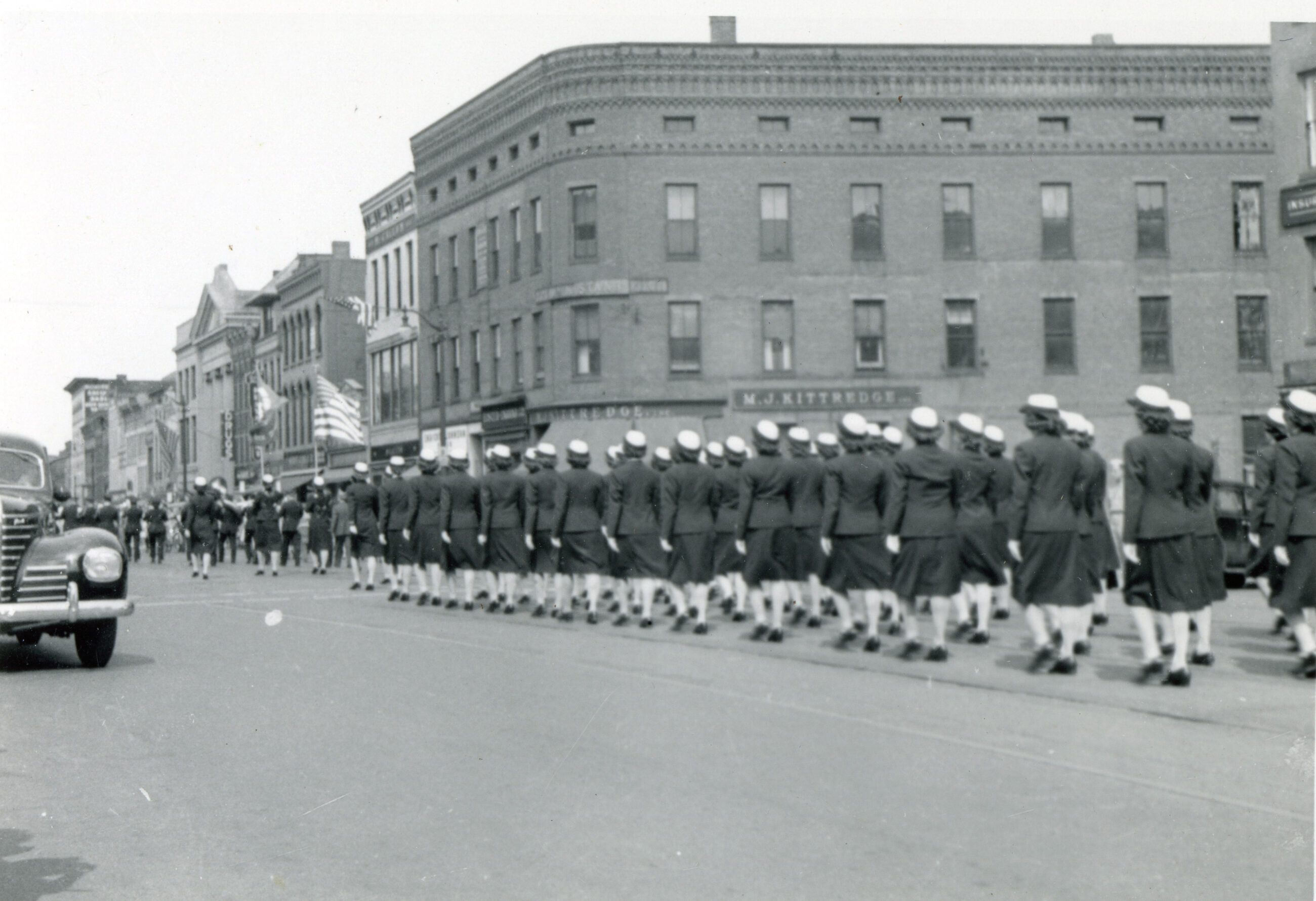Women in uniform (jackets and skirts) march in formation down Main Street.