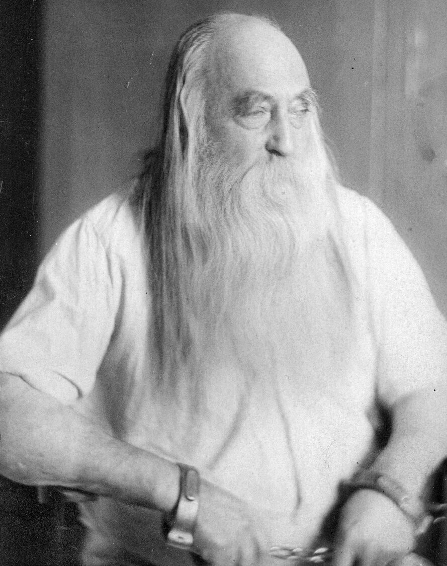 A black and white photograph of an elderly man with a very long, flowing white beard and long white hair parted down the middle. He is looking off to his right. He wears a loose-fitting white shirt, and his wrists are held together by shackles.