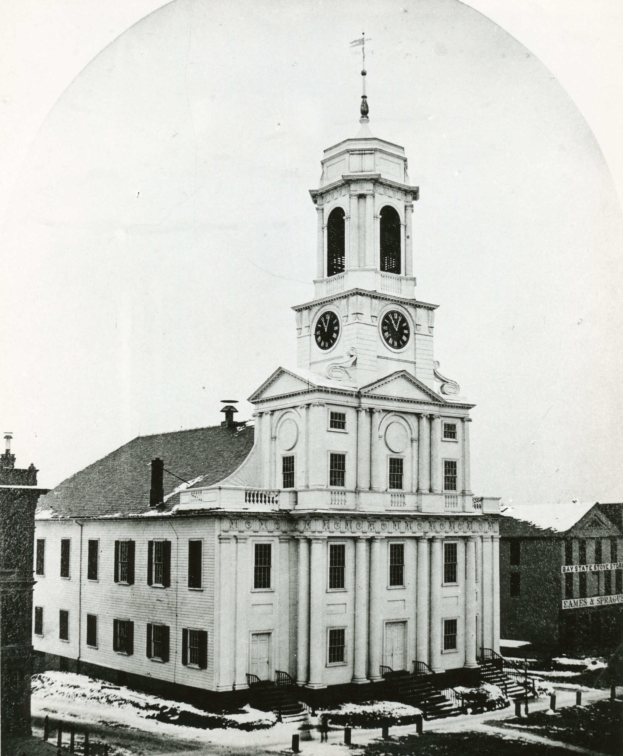 A simple white building with a bell tower and clock at the front, and columns around the entrance.