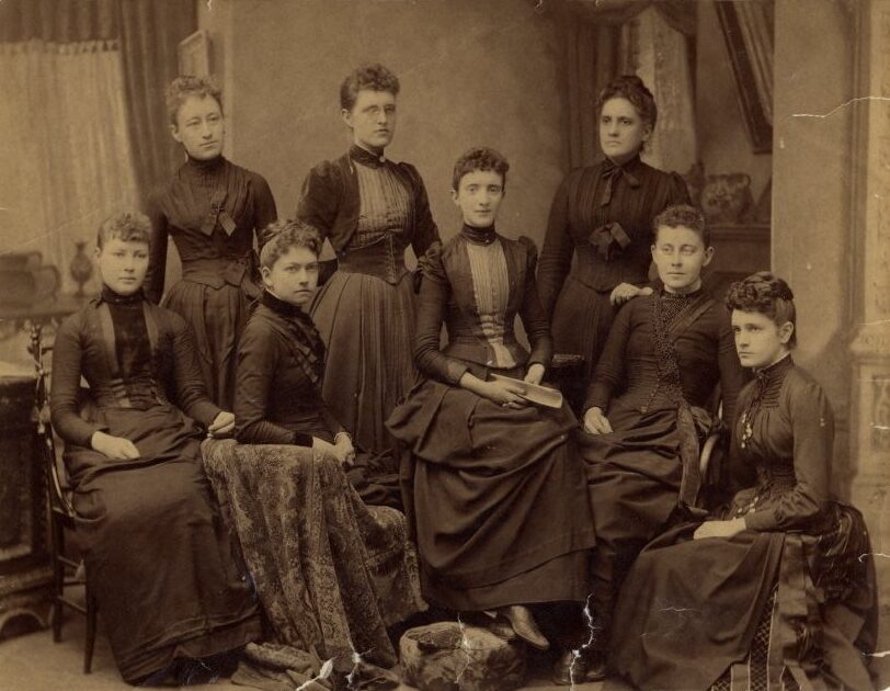 Group portrait of eight woman in a parlor setting.
