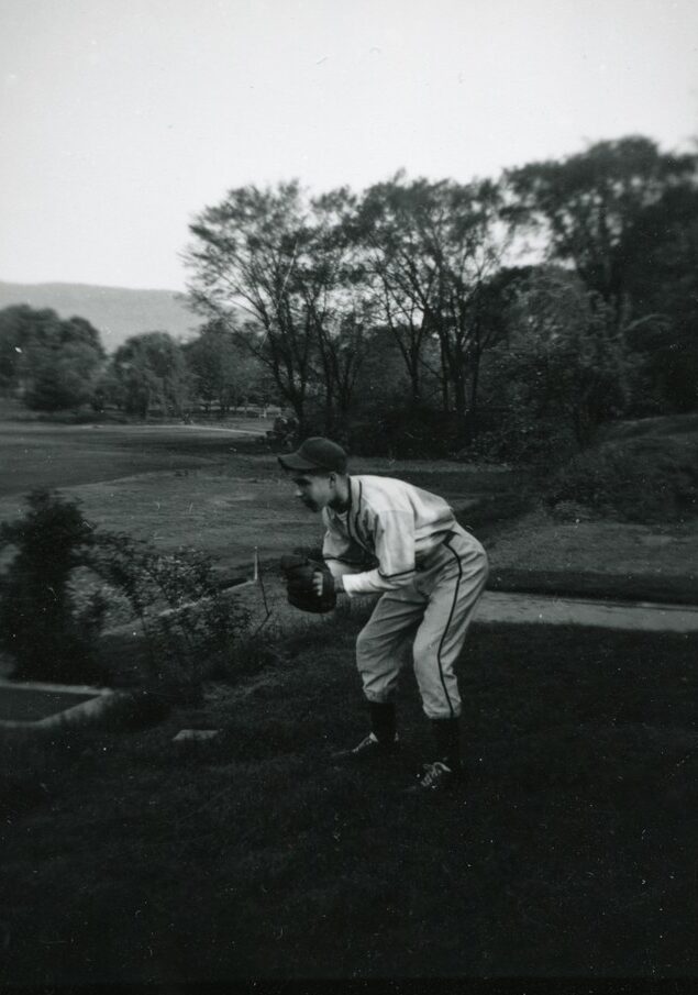 Black-and-white photograph of a teenaged baseball in uniform, posed crouching, with a baseball glove. There is a grassy field, trees, and a distant mountain in the background.