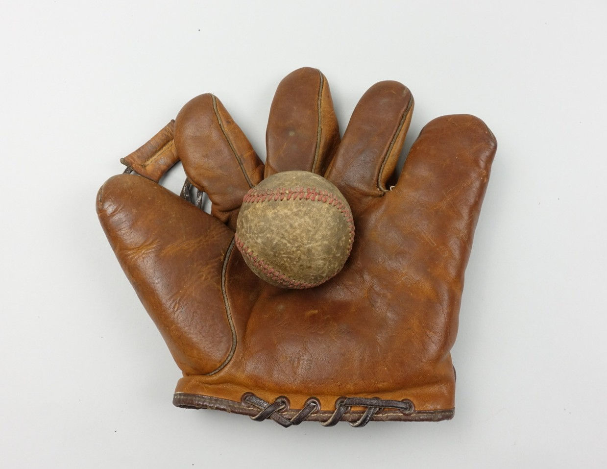 Photograph of an old, discolored white baseball with red stitching in the palm of an old, leather baseball glove with separated fingers, with a piece of leather connecting the index finger and thumb. 