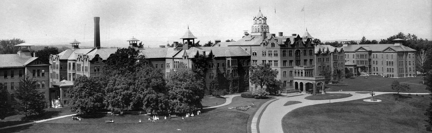 Black-and-white panoramic photograph of Northampton State Hospital showing the sprawling hospital campus with multiple connected brick buildings, a central tower, landscaped lawns, mature trees, curving pathways, and a smokestack rising above the complex.