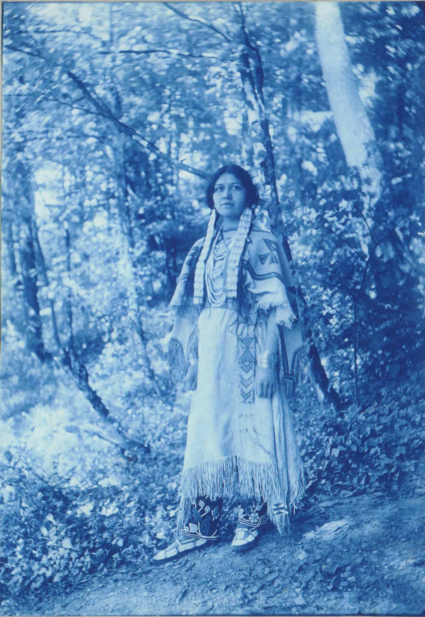Black-and-white photograph that appears blueish of Angel de Cora. Her hair is in two braids and she wears traditional Indigenous clothing. Her dress is light-colored and made of buckskin or cloth. It has long, dense fringe running along the bottom hemline and edges. The background is a wooded area.