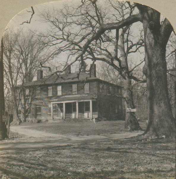 Black-and-white photograph of large two-story wooden building with a front porch, multiple chimneys. A dirt circular drive leads up to the house. Surrounded by woods