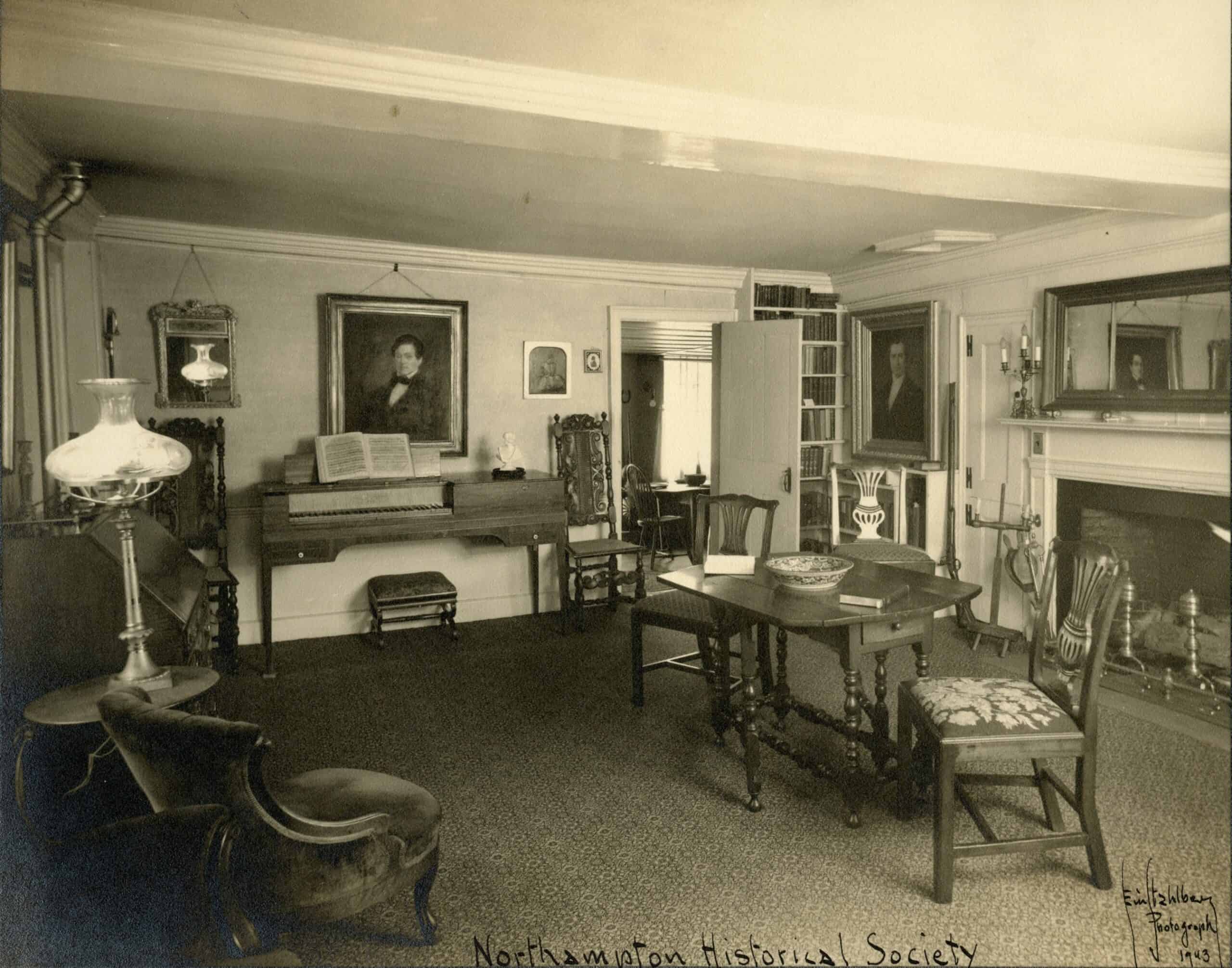 A black-and-white photograph shows an interior parlor filled with antique furnishings and decor, documented by the Northampton Historical Society in 1943. In the left foreground, a tufted armchair faces toward the center of the room, positioned next to a small round side table holding a tall, ornate glass oil lamp with a wide, frosted shade. Behind it sits a dark wood slanted-front writing desk. Along the back-left wall, a square clavichord or spinet piano stands with an open music book resting on its rack, paired with a small upholstered footstool underneath. Hanging directly above the instrument is a large, dark-framed portrait of a man, flanked by a smaller decorative mirror to its left and miniature framed portraits to its right. Two tall, ornately carved wooden chairs flank the musical setup. An open doorway near the center of the background reveals a glimpse into an adjacent, sunlit dining room containing a table and chairs.To the right of the doorway, a tall, built-in bookcase filled with books stands next to another large framed portrait hanging on the wall. In the center-right foreground, a wooden drop-leaf table features turned legs and holds a wide ceramic bowl and a book. It is surrounded by three matching wooden chairs with patterned, upholstered seats. On the far right, there is a large brick fireplace with a white mantelpiece, adorned with a long, horizontal rectangular mirror hanging above it and a pair of candlesticks on the shelf. A spinning wheel sits tucked in the corner between the bookcase and the fireplace hearth. The entire room is covered in a dark, textured wall-to-wall carpet. Handwritten text along the bottom border reads "Northampton Historical Society" near the center, and a photographer's signature with the date "1943" is visible in the bottom right corner.