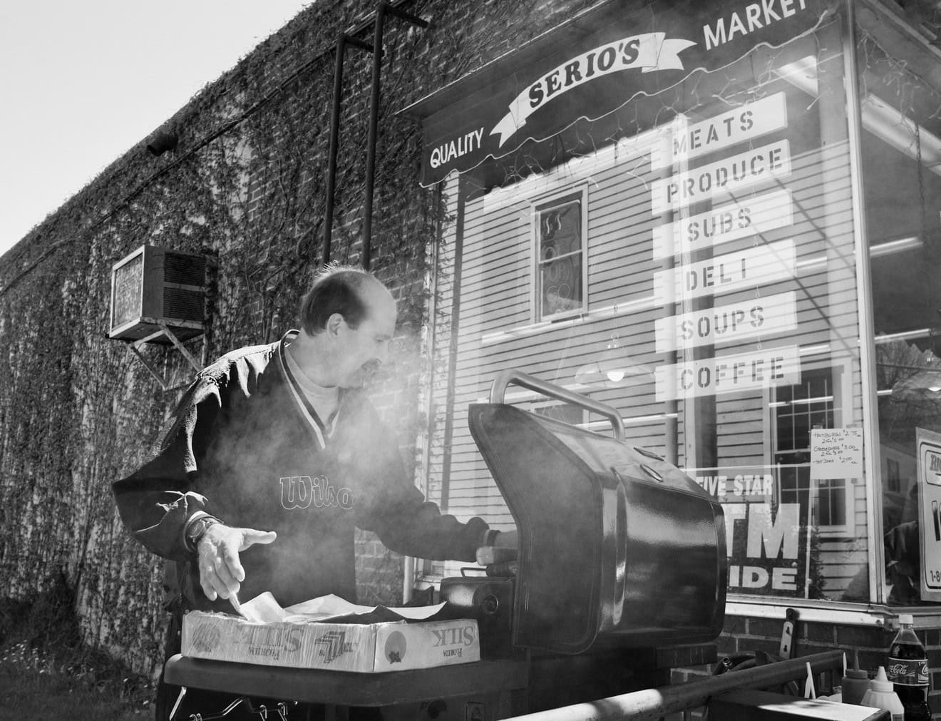 Black-and-white photograph of a middle aged man with dark balding hair and a mustache cooking something on the a barbeque grill. His face is blurred by smoke. Behind him is a storefront with a sign that reads: "QUALITY SERIO'S MARKET."
