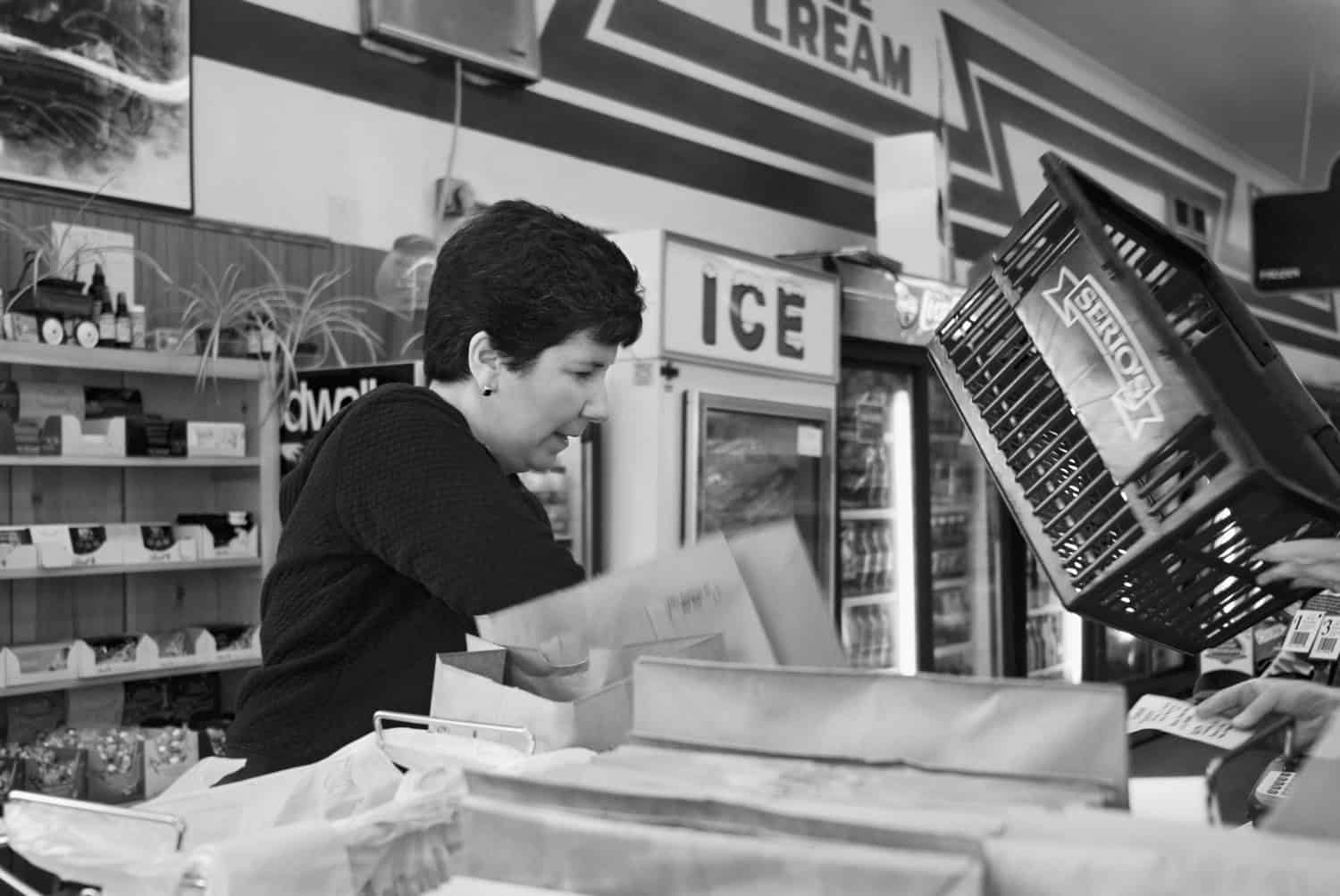 Black-and-white photograph of a middle aged woman with dark short hair and a dark top working at a cluttered convenient store counter. An ice machine and refrigerators full of beverages are seen in the distance.