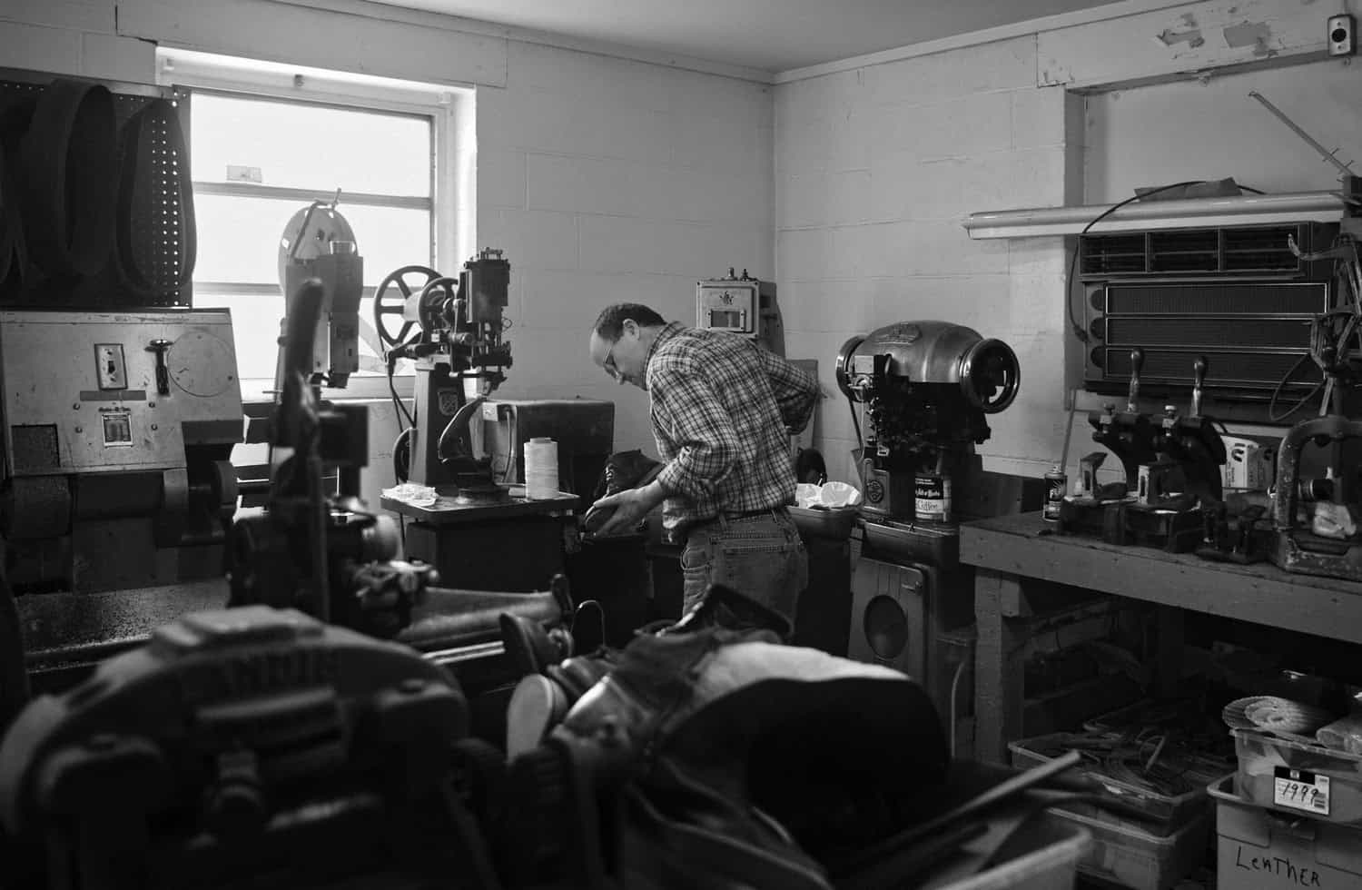Black-and-white photograph of a man in an office or workshop examining a black boot. He is faced away from the camera and has dark short hair. He is wearing a plaid shirt tucked into jeans.