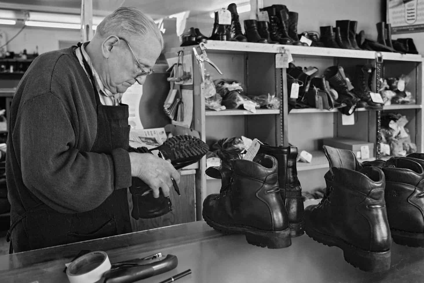 Black-and-white photograph of a matured man looking at the bottom of a black boot. He has on a longsleeve sweatshirt and a black apron. Behind him are shelves of black boots.