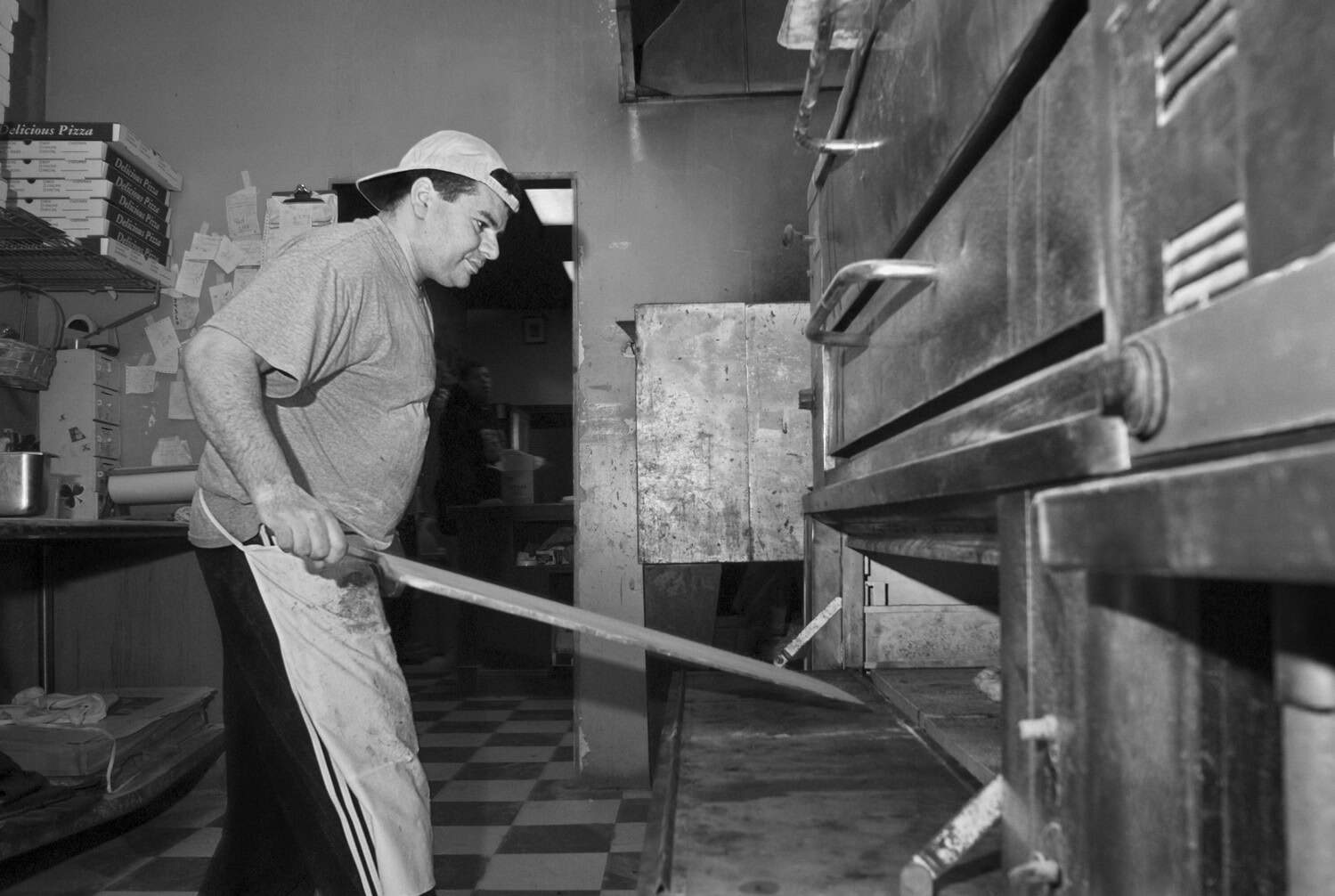 Black-and-white photograph of a man working at a pizza shop, flipping cooking pizza or taking it out of the oven. He is wearing a t-shirt and has his white apron tied around his waist. He has on a backwards baseball hat.