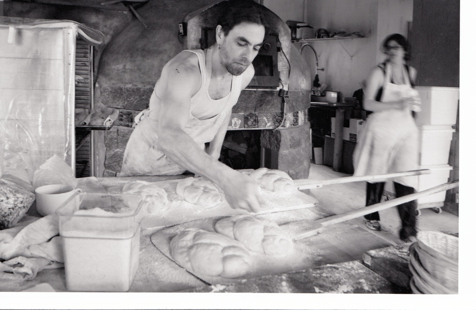 Black-and-white photograph of a man in a white tank top and apron preparing dough to be put in the oven. A woman with an apron on can be seen walking toward the camera in the distance. They are in a kitchen and industrial stoves can be seen in the background.