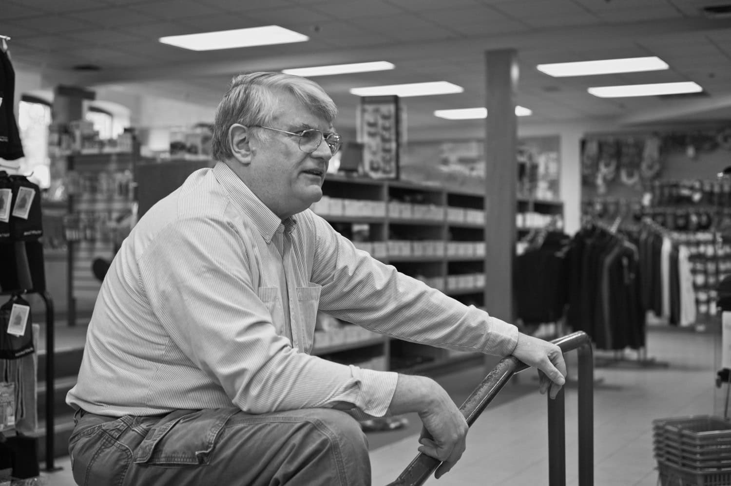 Black-and-white photograph of a middle aged man sitting and talking to someone off to the side past the camera. He has white hair and glasses and is wearing a buttoned down shirt. Shelves can be seen behind him.