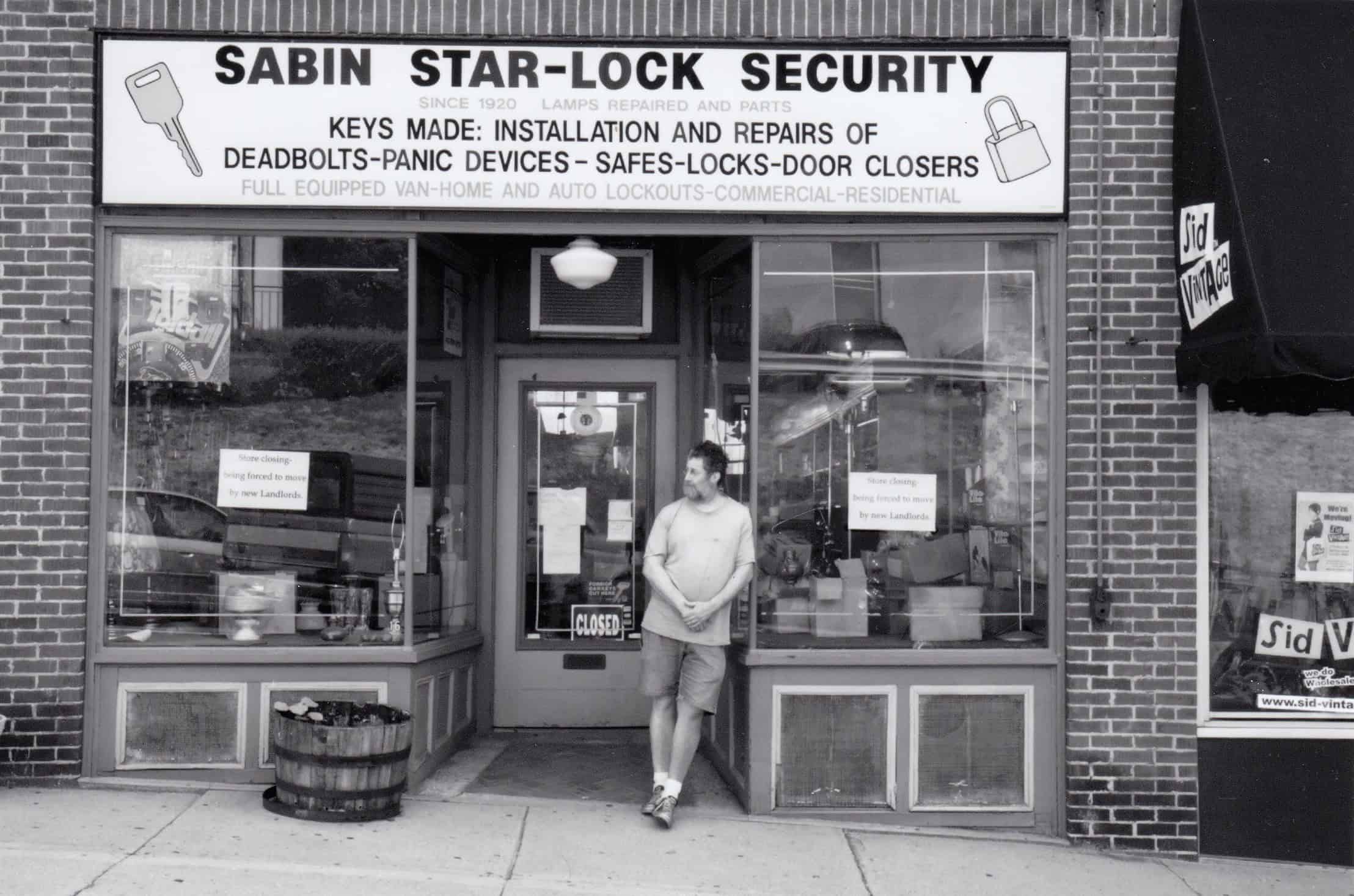 Black-and-white photograph of a man leaning against a storefront. The sign of the store is above the door and reads: "SABIN STAR-LOCK SECURITY KEYS MADE: INSTALLATION AND REPAIRS OF DEADBOLTS-PANIC DEVICES-SAFES-LOCKS-DOOR CLOSURES." The main has dark short hair and is bearded. He is wearing a t-shirt, shorts, and sneakers.