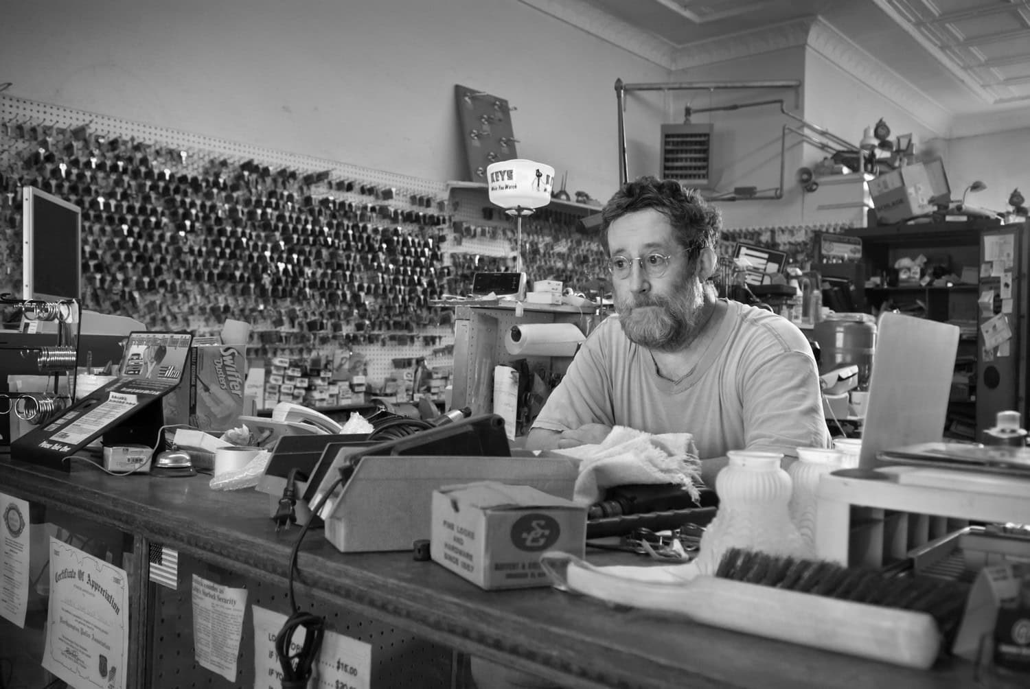 Black-and-white photograph of a middle aged man with dark graying hair and glasses sitting at a checkout counter. Behind him is a wall full of hanging keys.
