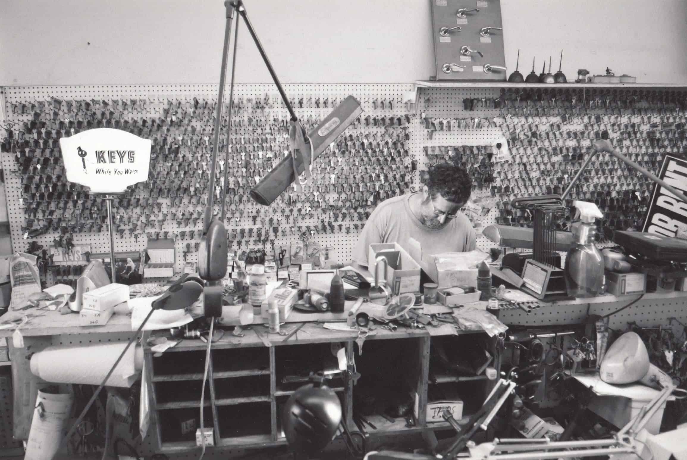 Black-and-white photograph of a man working at a cluttered desk. Behind him is a wall full of hanging keys.